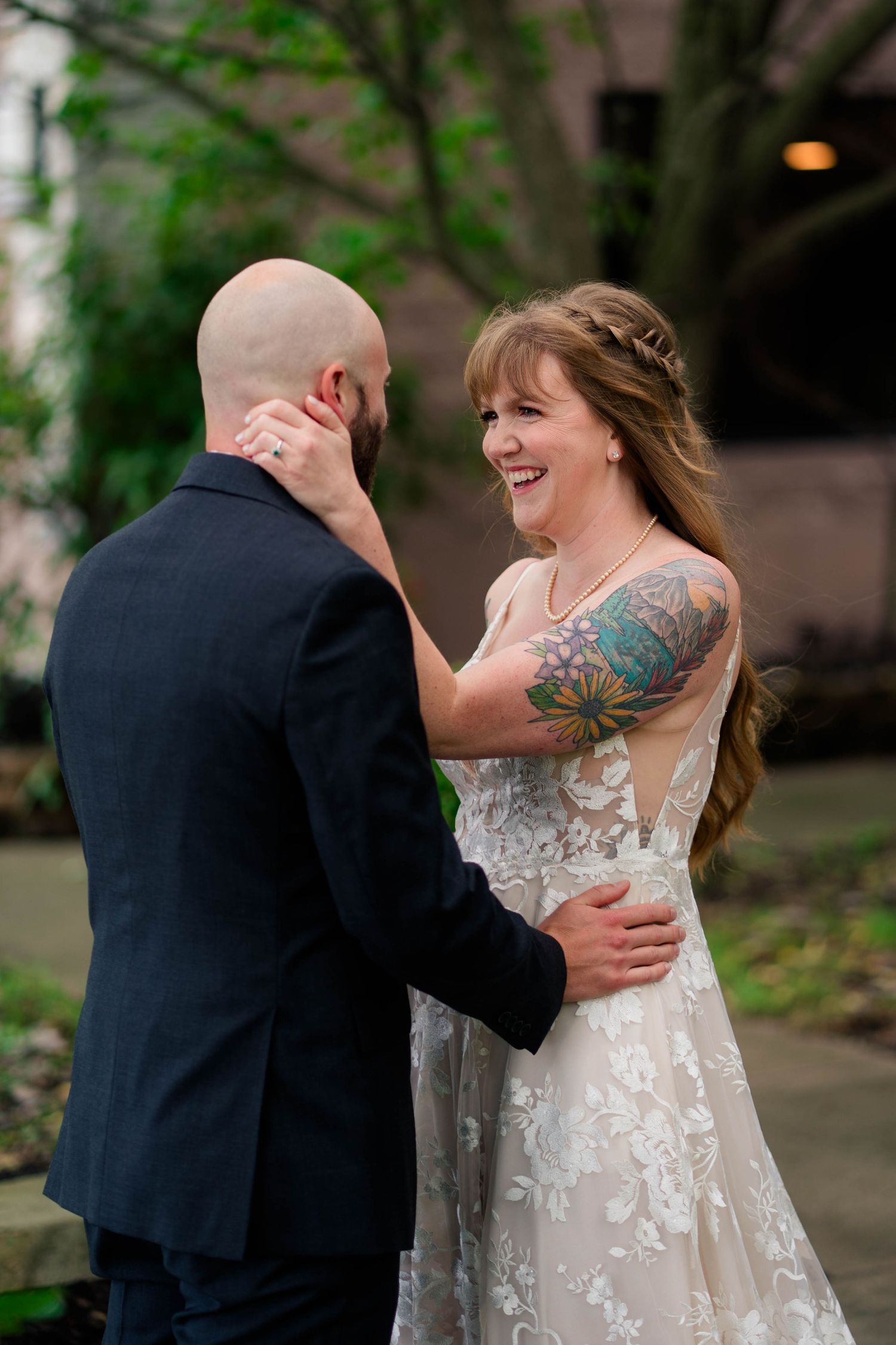 Tattooed bride laughs joyfully while embracing bald groom during outdoor wedding first look.