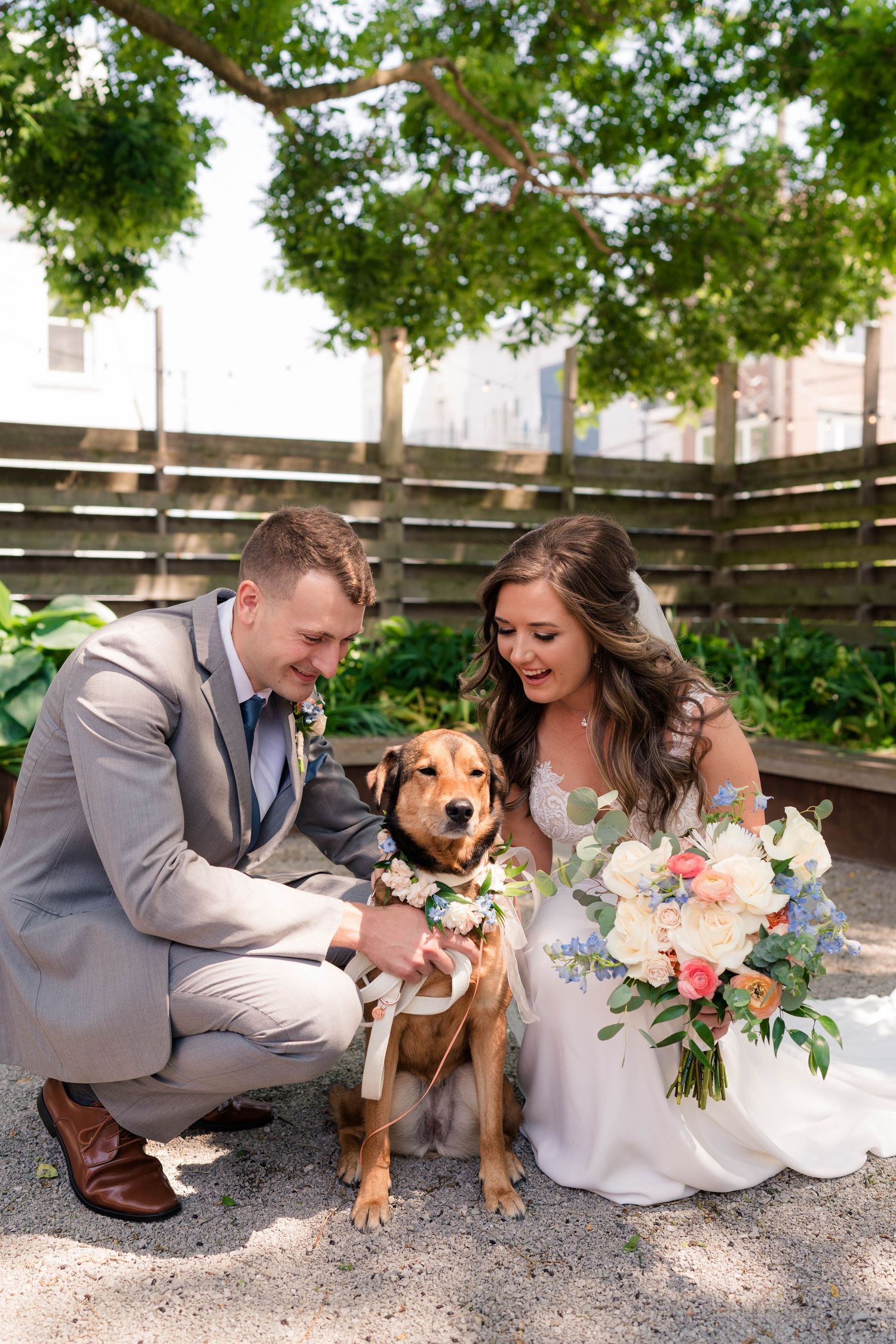 Bride and groom crouch beside their flower-crowned dog during outdoor wedding portraits.