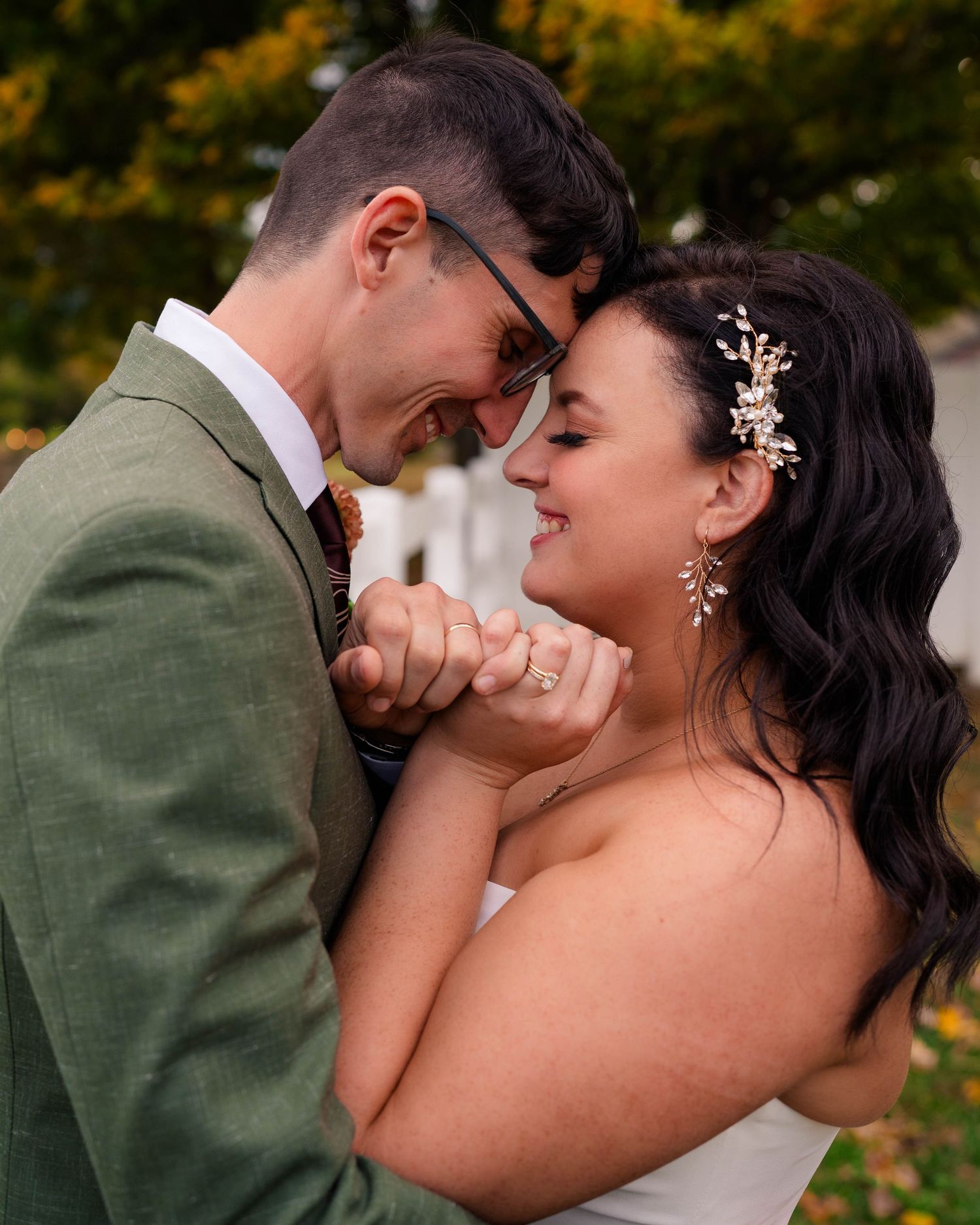 Bride and groom touch foreheads and smile while holding hands outdoors in autumn setting.