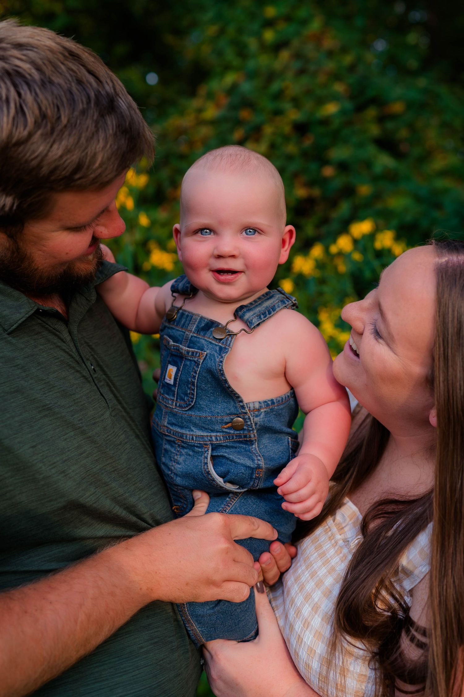 Happy baby in denim overalls smiling at parents while being held outdoors near yellow flowers.
