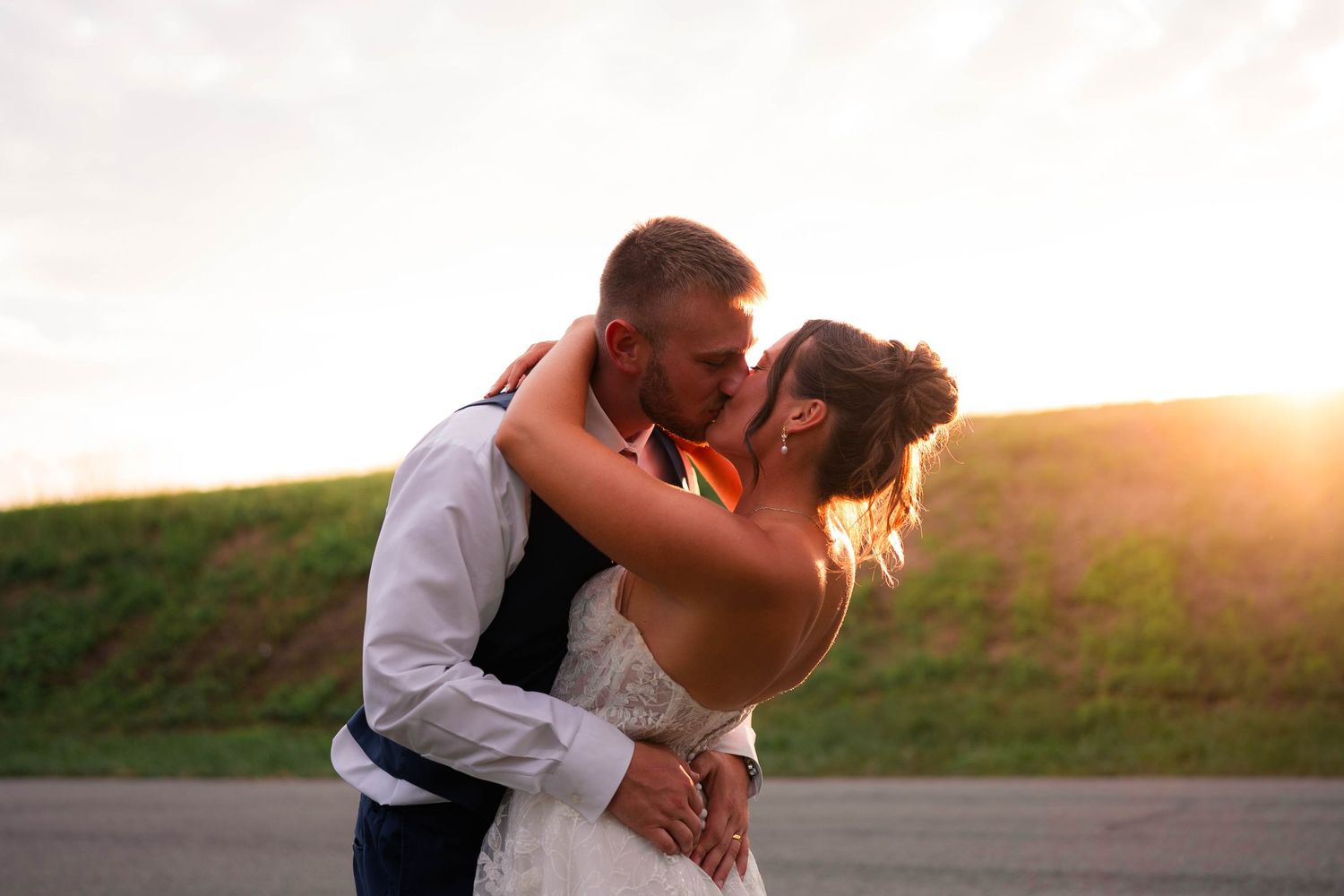 Newlyweds share a romantic kiss outdoors with a glowing golden sunset backdrop on their wedding day.