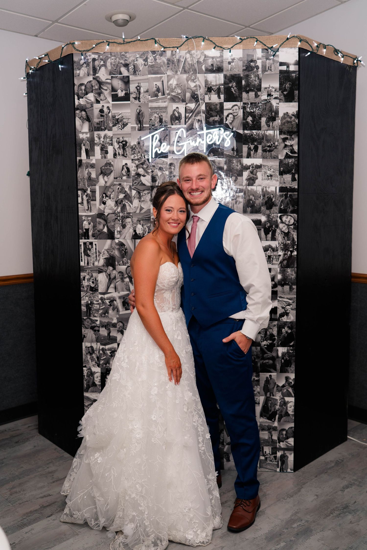 Bride and groom pose together in front of a black and white photo collage backdrop with neon sign at wedding reception.