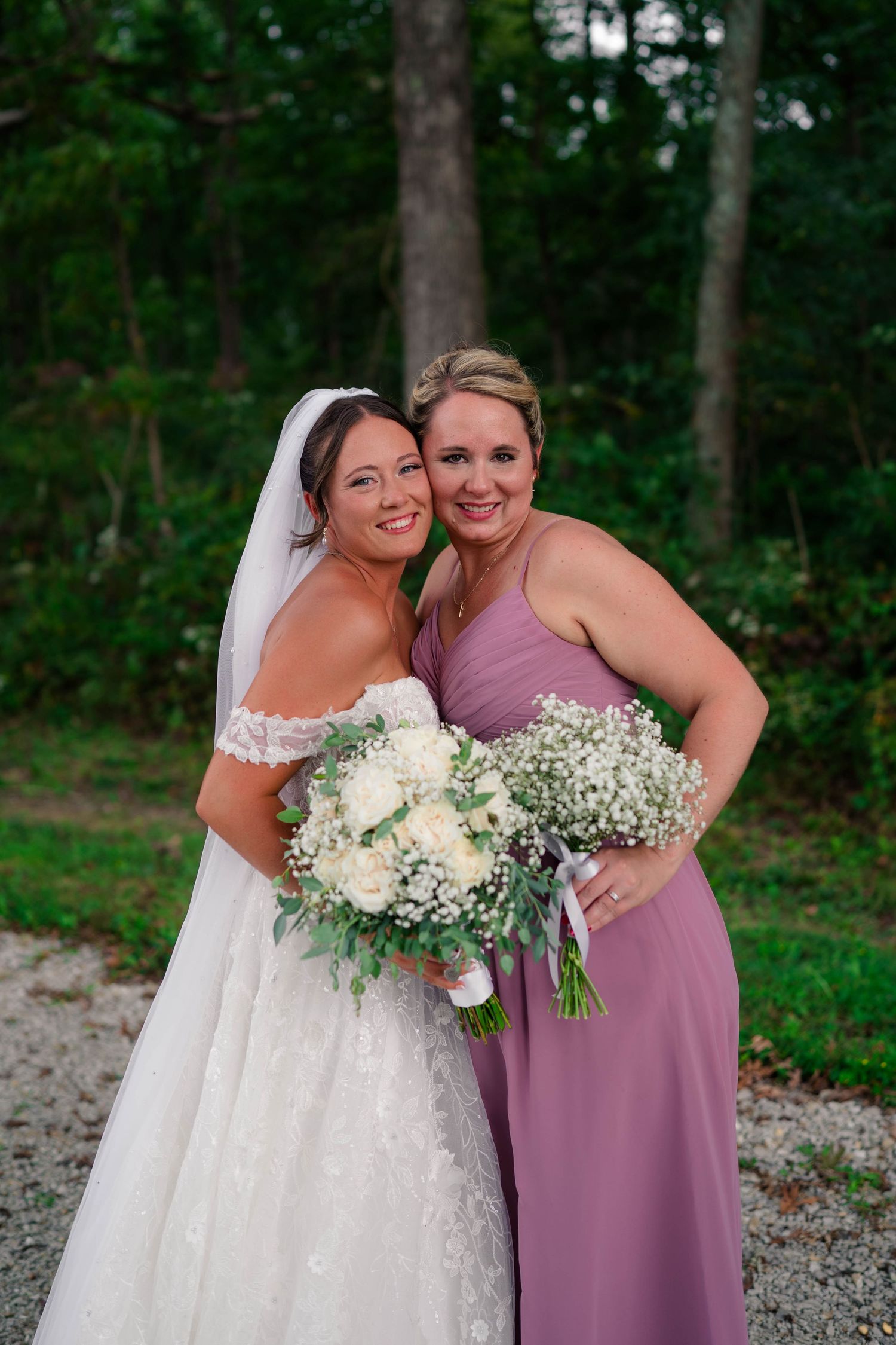 Bride in lace wedding gown and bridesmaid in mauve dress pose together holding white and baby's breath bouquets.