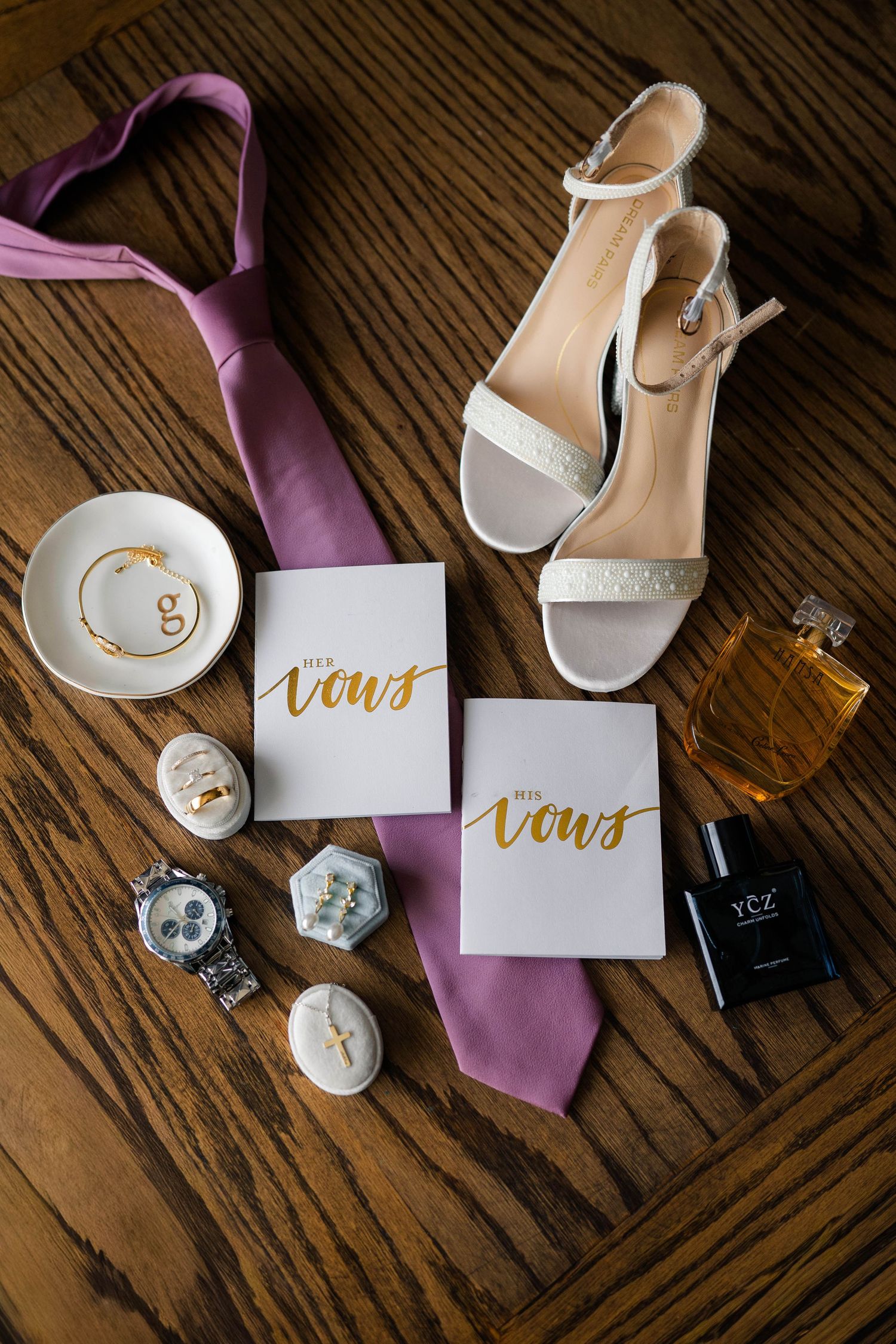 Flat lay of wedding accessories including vows books, heels, ties, rings, watch, and perfume on wood.