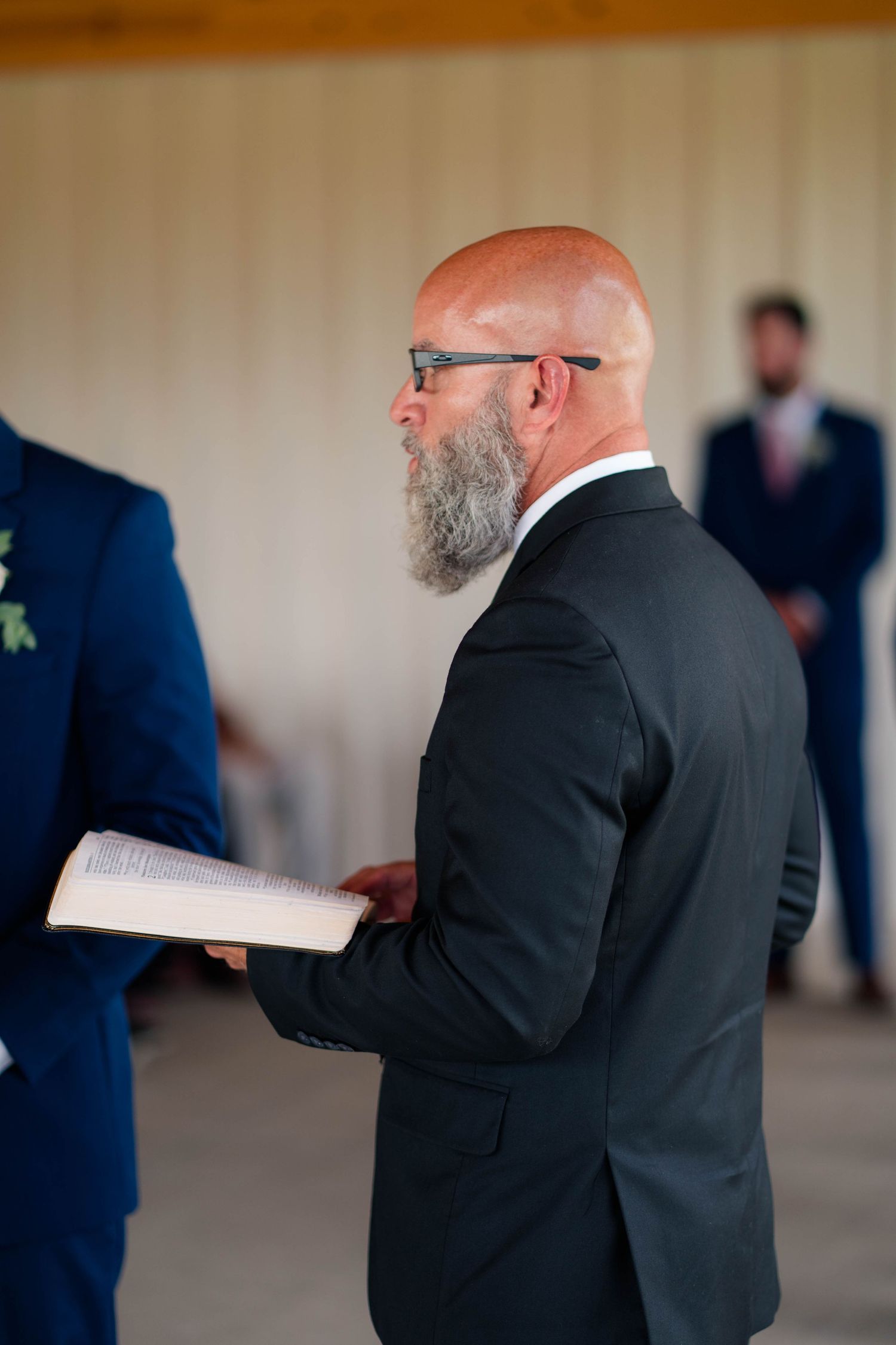 Bald officiant with gray beard reads from book during wedding ceremony wearing black suit and glasses.