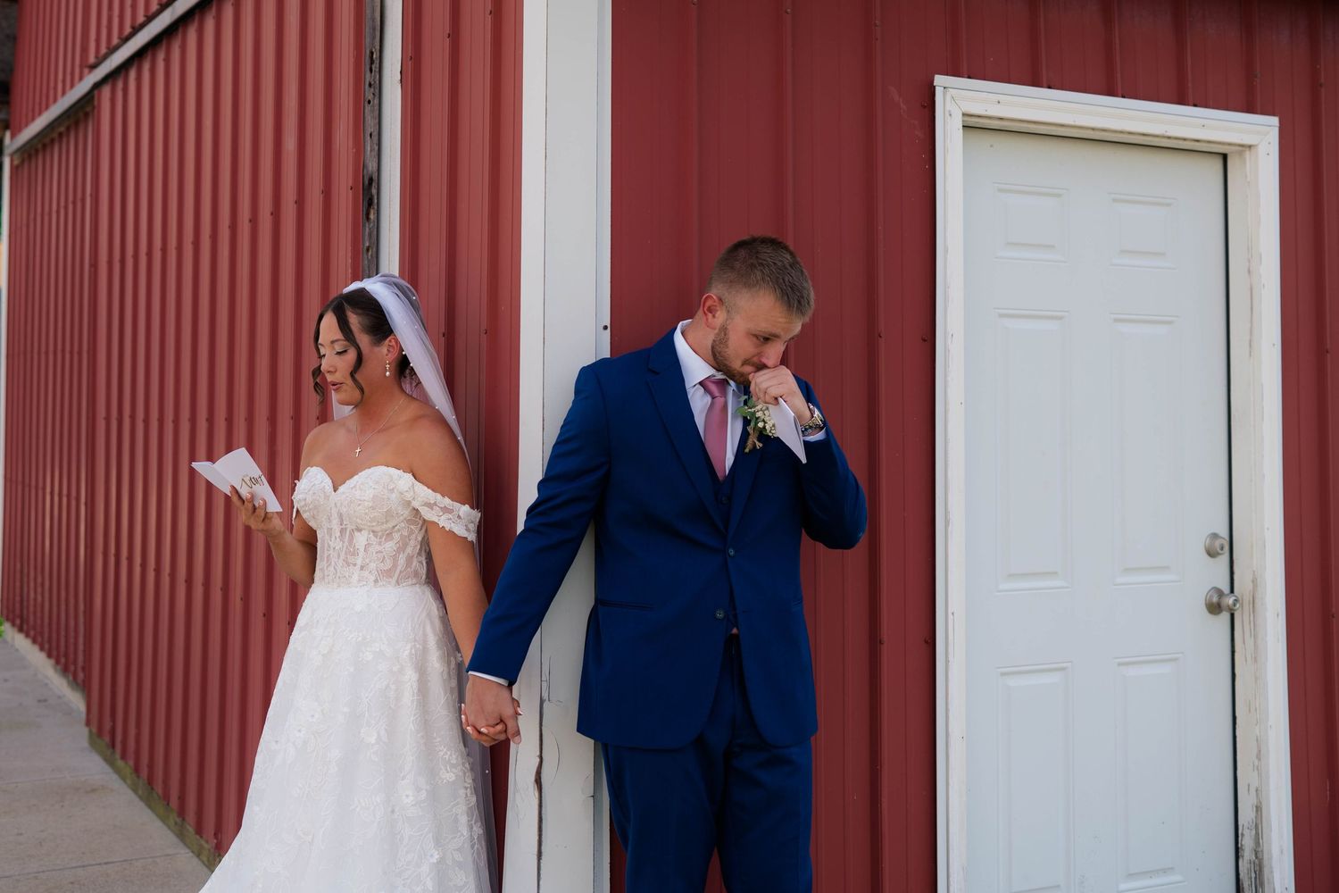 Bride and groom share emotional first look moment outside red barn, reading letters before wedding ceremony.