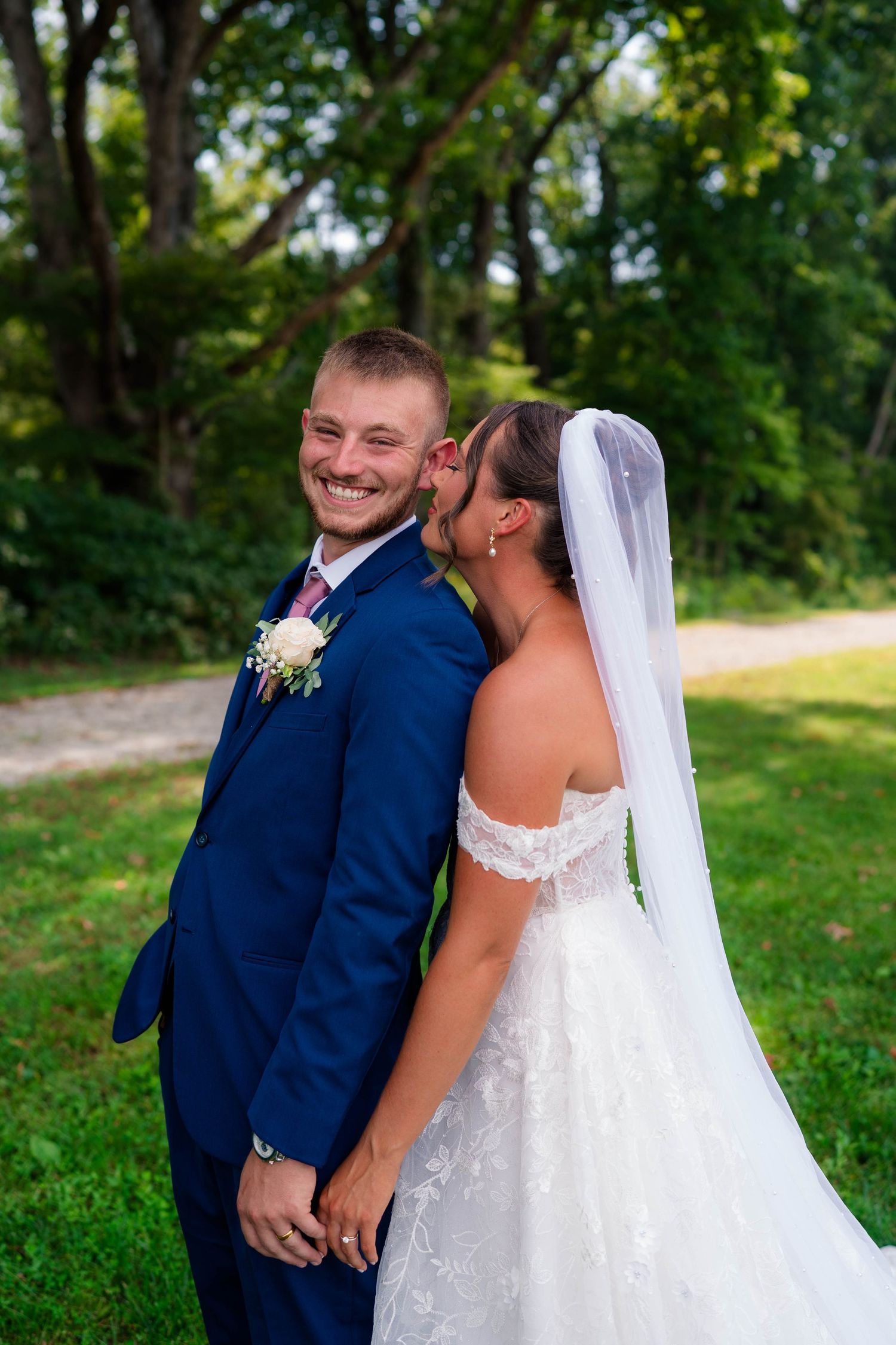 Smiling groom in navy suit holds bride's hand as she kisses his cheek outdoors on their wedding day.