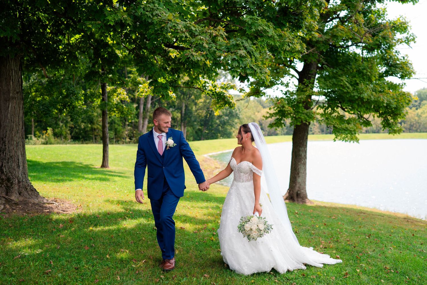 Bride and groom hold hands walking on green lawn near a serene lake on their wedding day.
