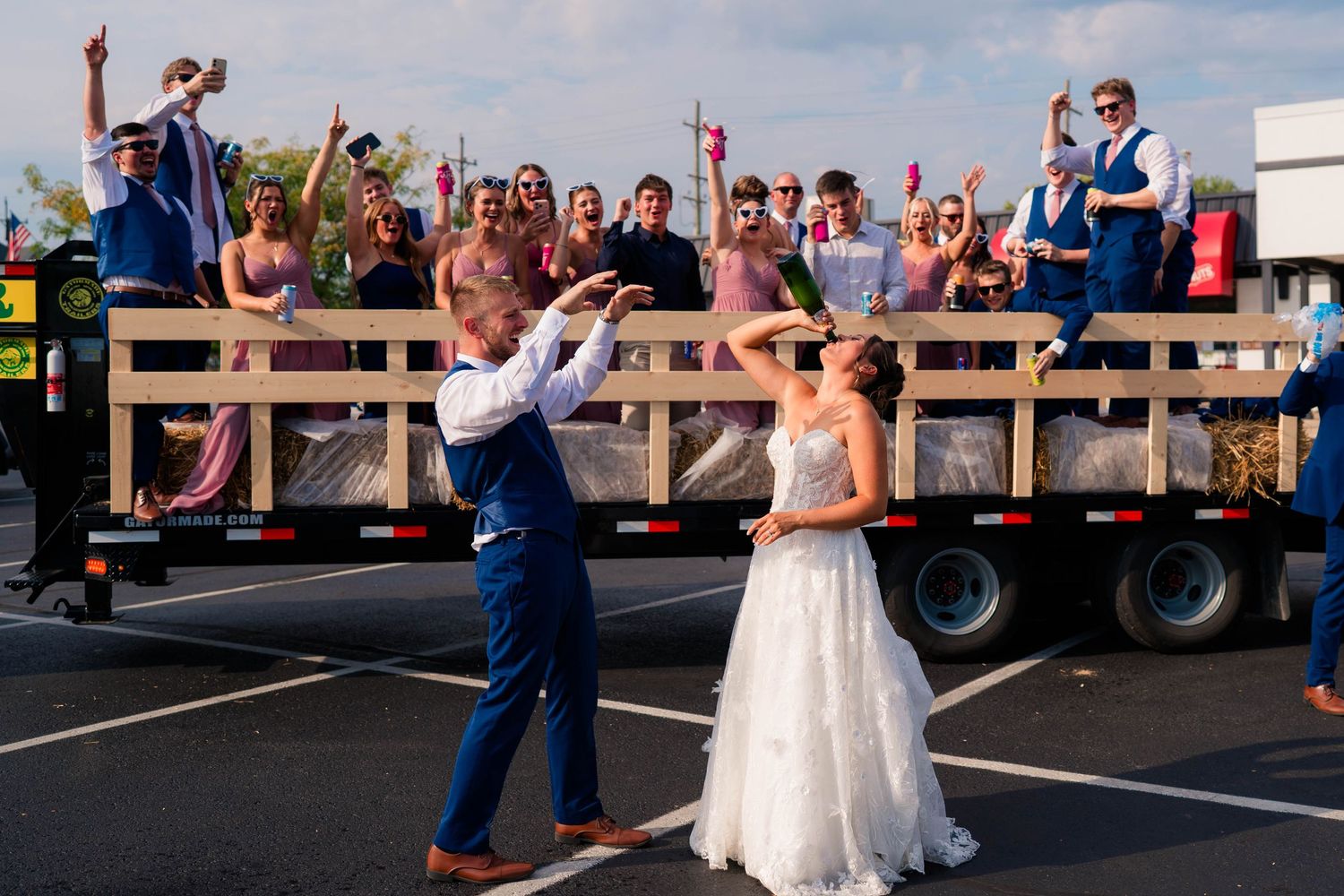 Bride and groom popping champagne in parking lot with wedding guests cheering on a flatbed.