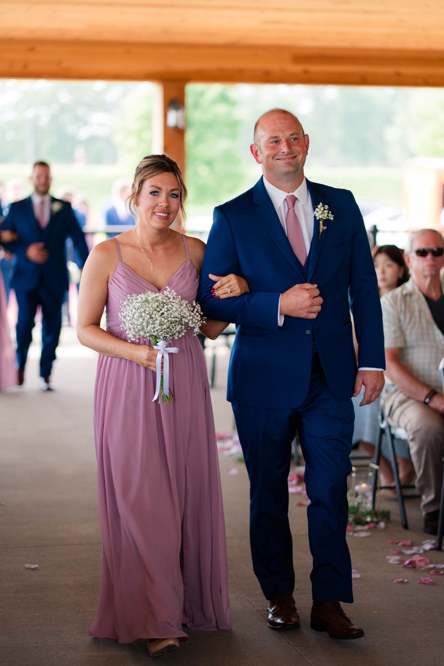 Bridesmaid in mauve gown with baby's breath bouquet walks down aisle with groomsman.