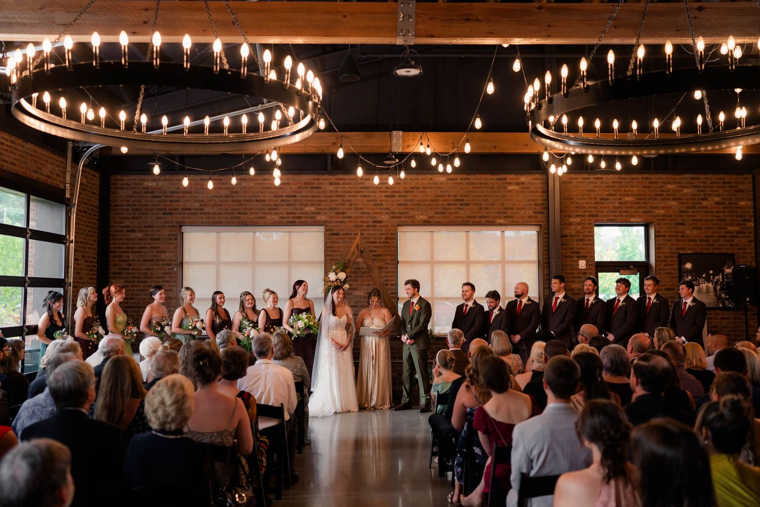Wedding ceremony in rustic brick venue with circular Edison bulb chandeliers overhead.