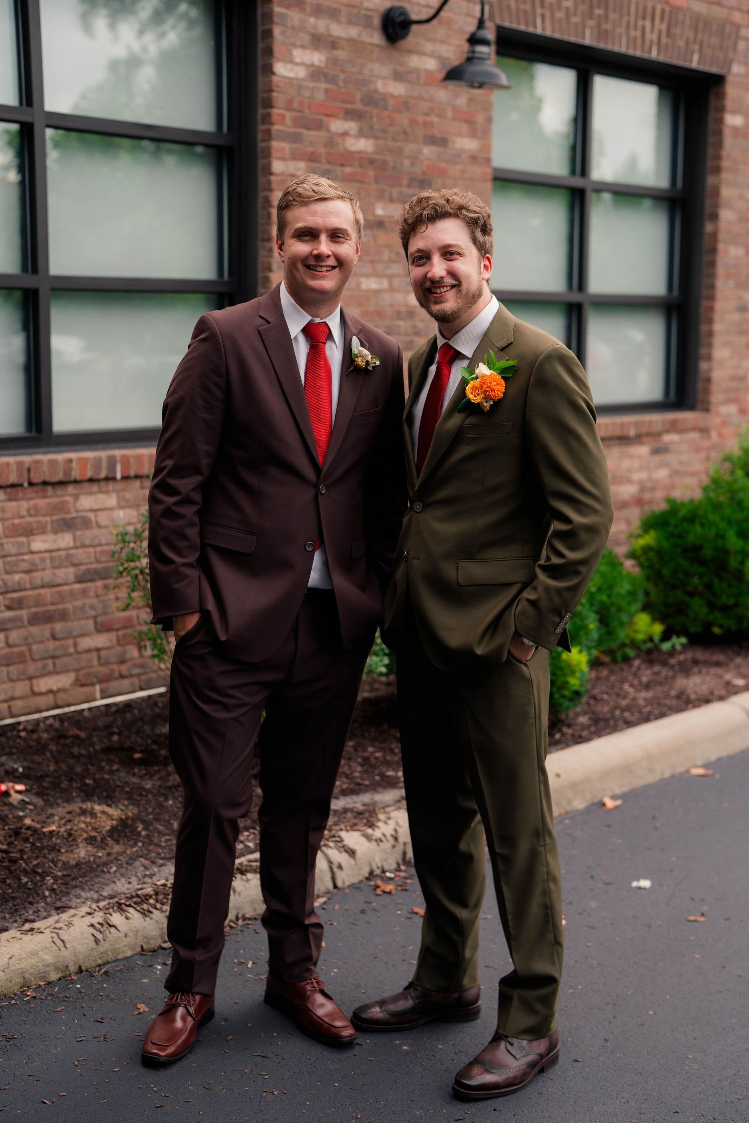 Two groomsmen in brown and olive suits with orange boutonnieres posing outside brick building.