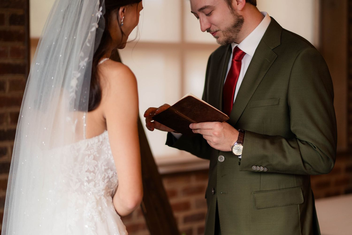 Groom in olive suit reading wedding vows from a book to his bride in a lace wedding dress.