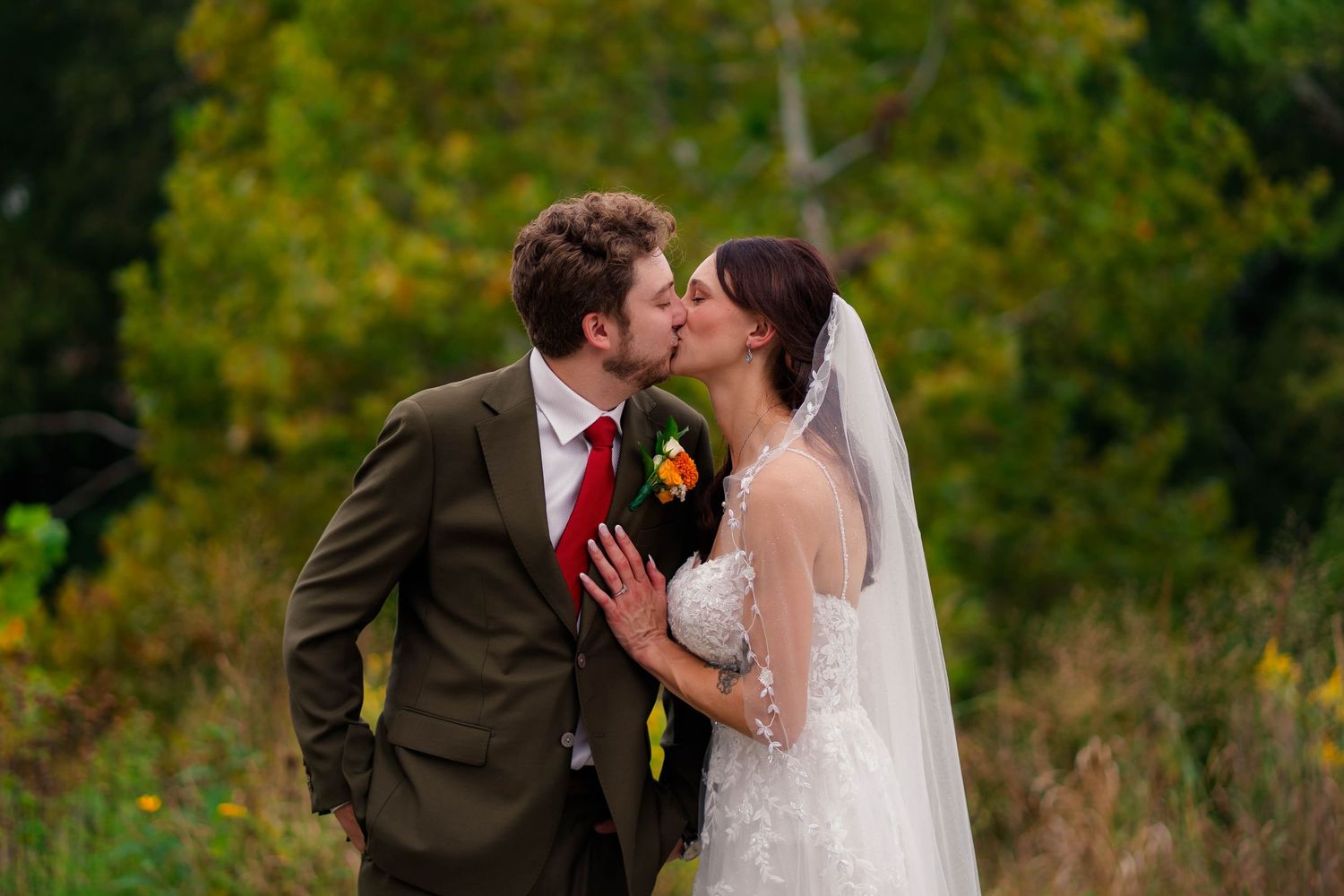 Bride and groom share a romantic kiss outdoors with colorful autumn foliage in the background.