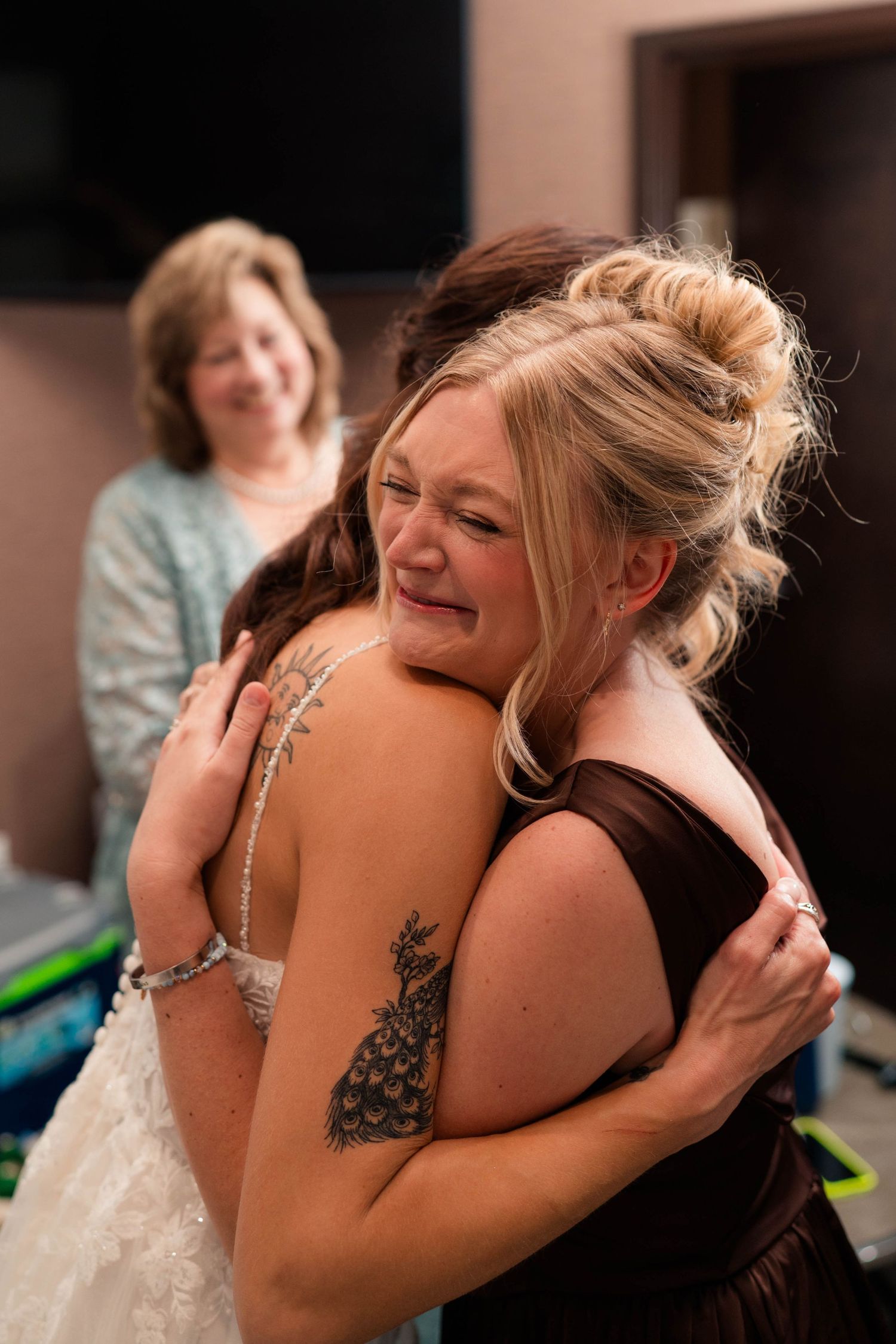 Emotional bride hugging bridesmaid in brown dress while woman smiles in background.