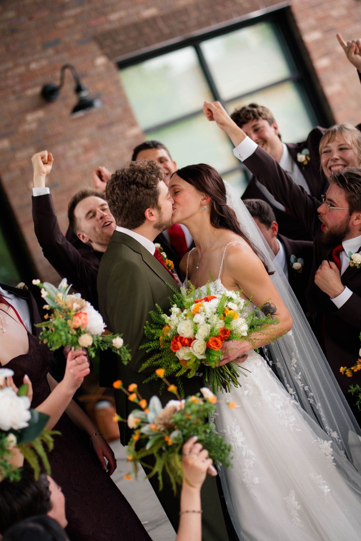 Bride and groom share a kiss surrounded by cheering wedding party holding colorful bouquets outdoors.