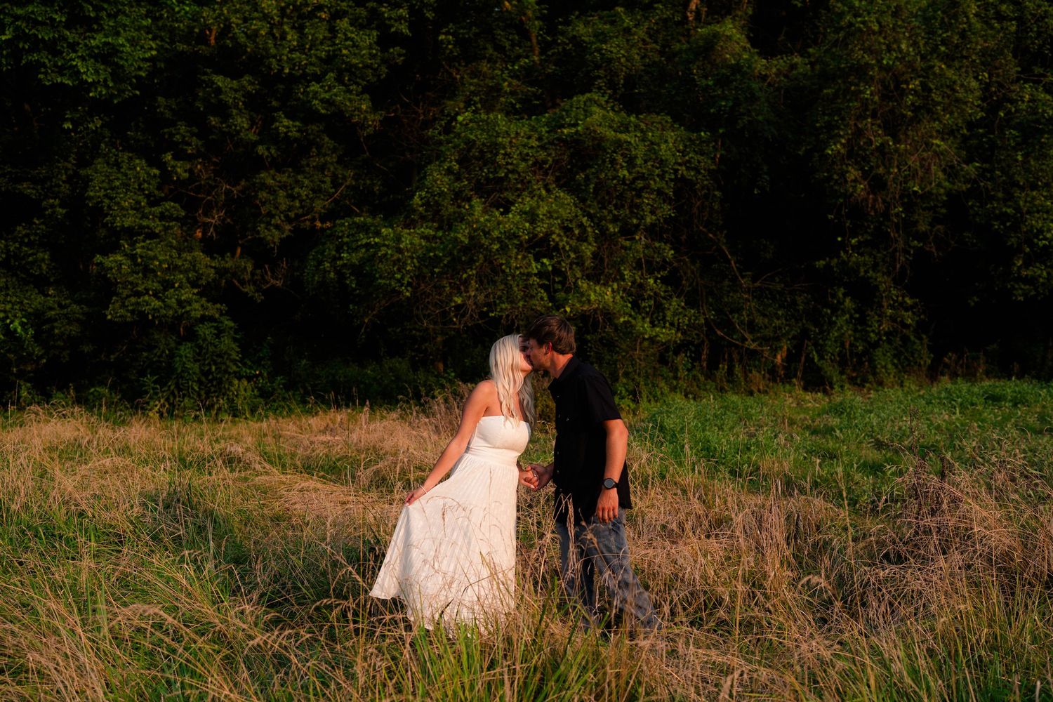 A bride and groom share a romantic kiss in a sunlit meadow with lush green trees in the background.