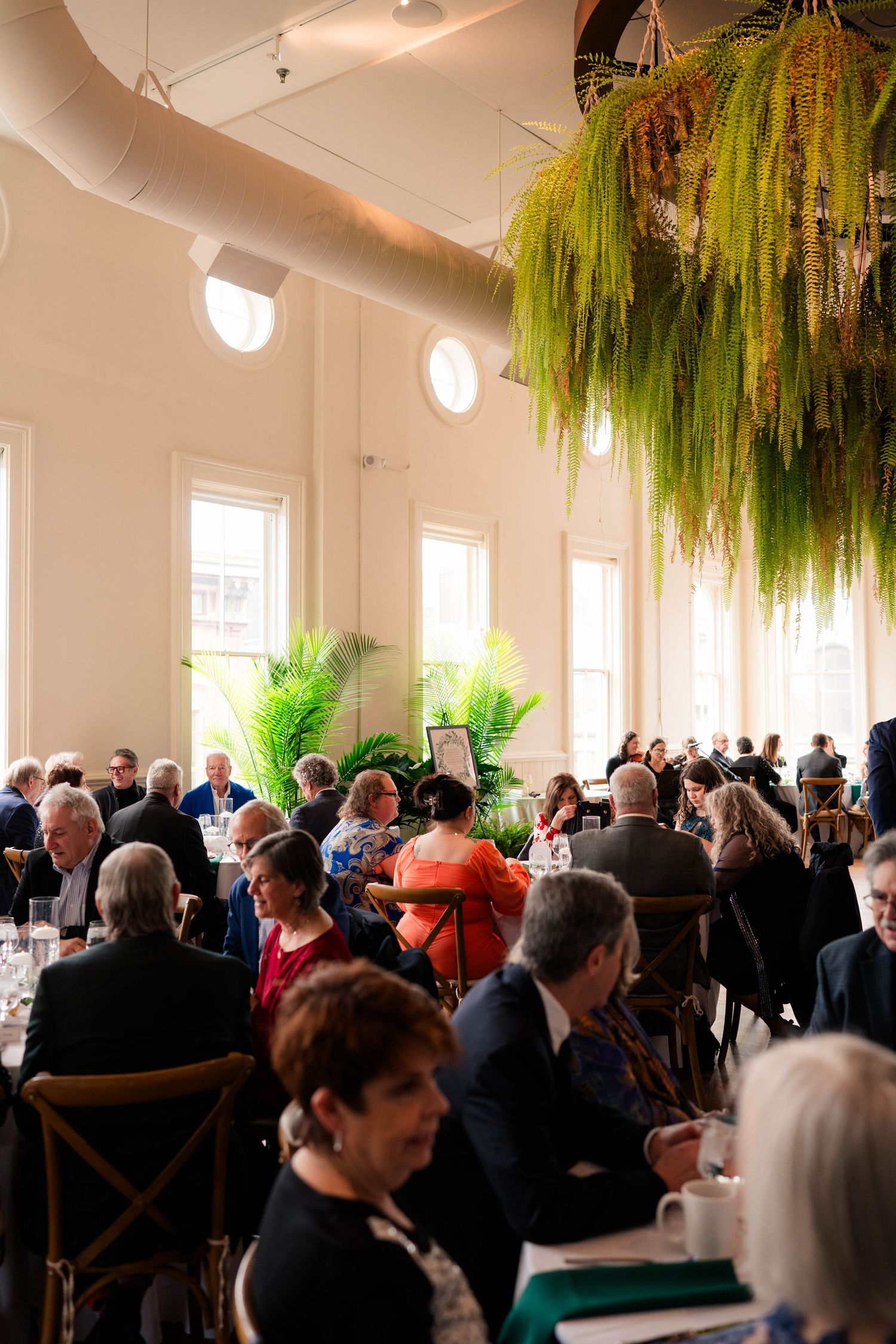 Crowded elegant event hall with high ceilings, hanging ferns, and guests seated at formal dining tables.