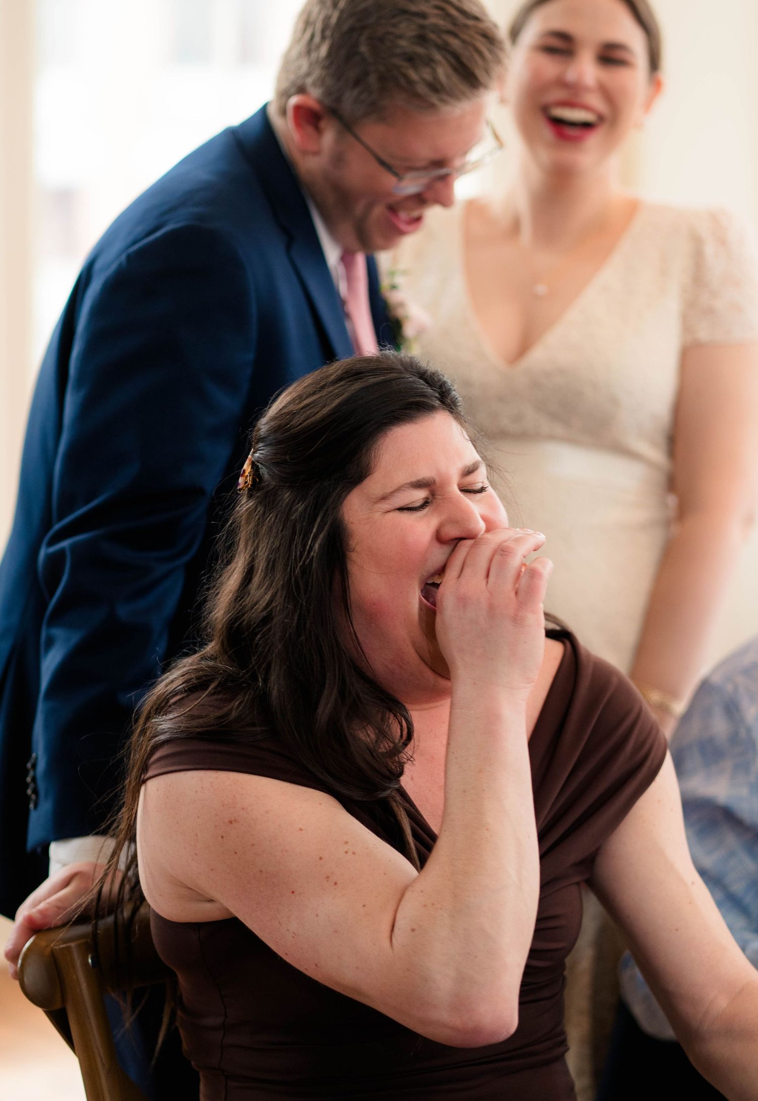 Woman laughing with hand over mouth at wedding reception as bride and groom smile behind her.