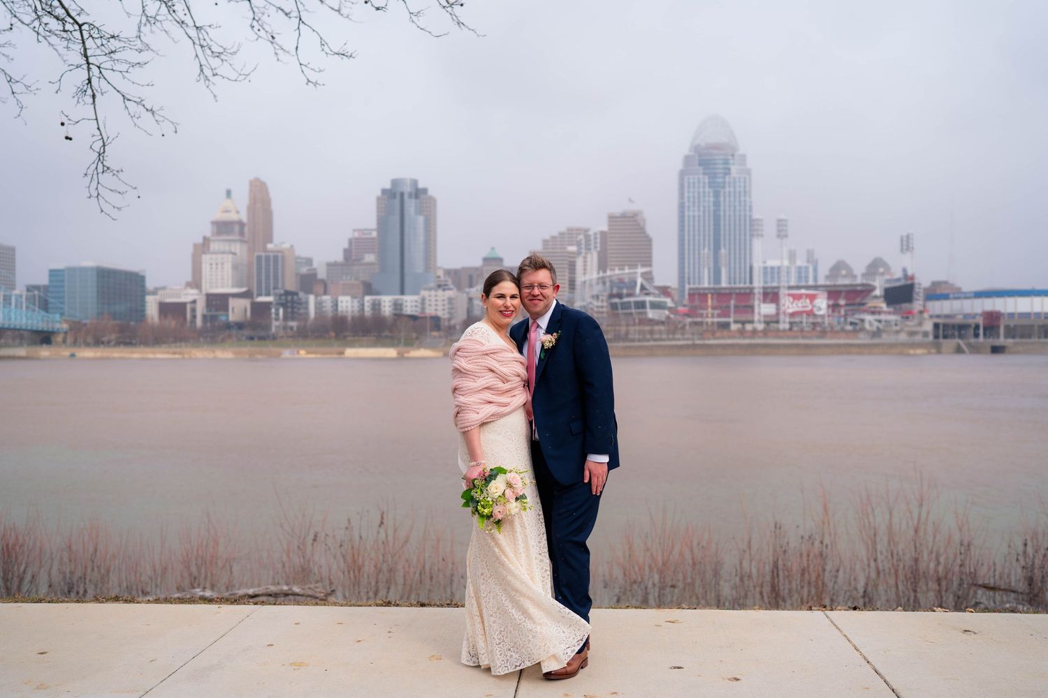 Bride and groom posing together by river with Cincinnati city skyline on overcast winter day.