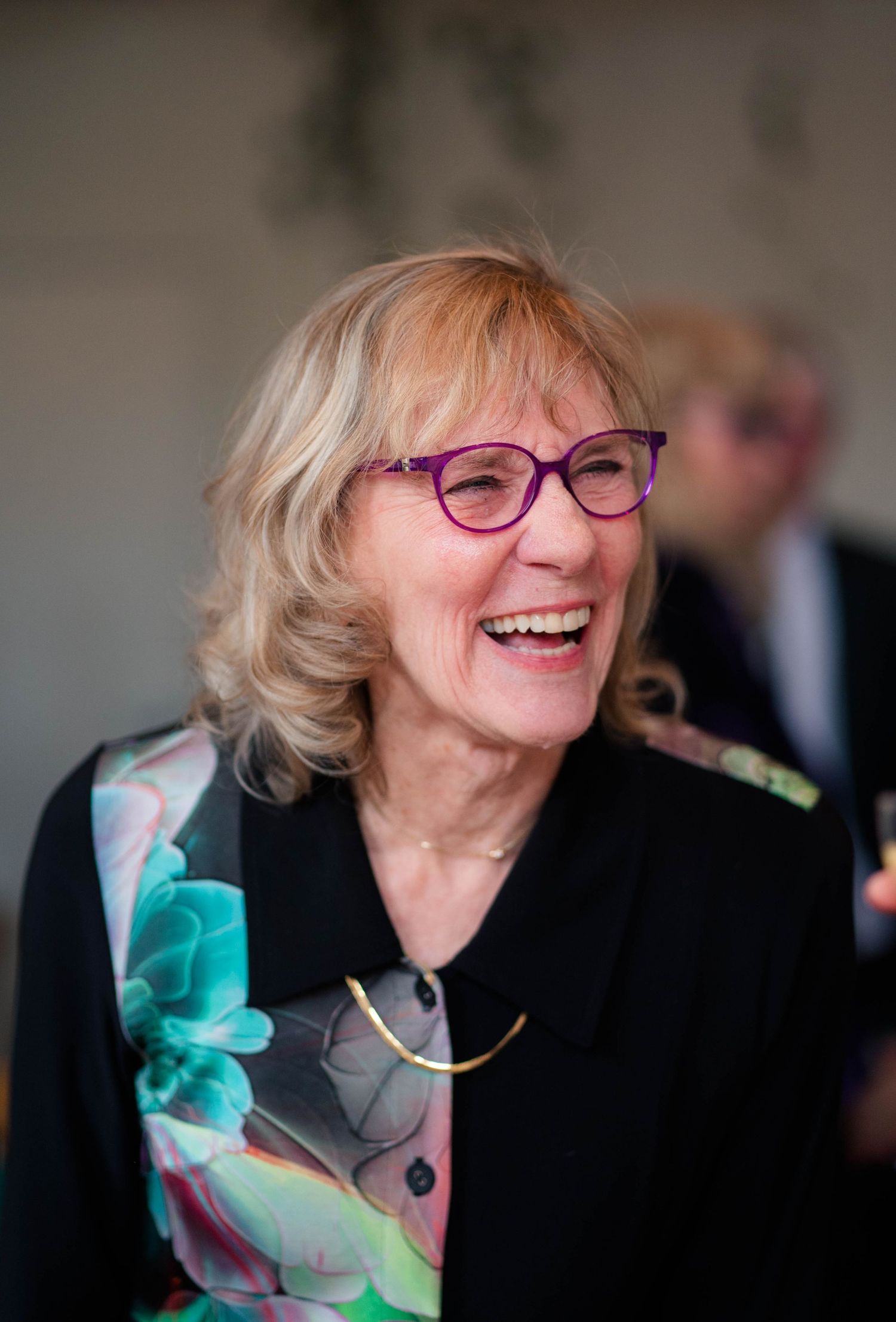 Joyful older woman with blonde hair and purple glasses laughing at a wedding celebration.