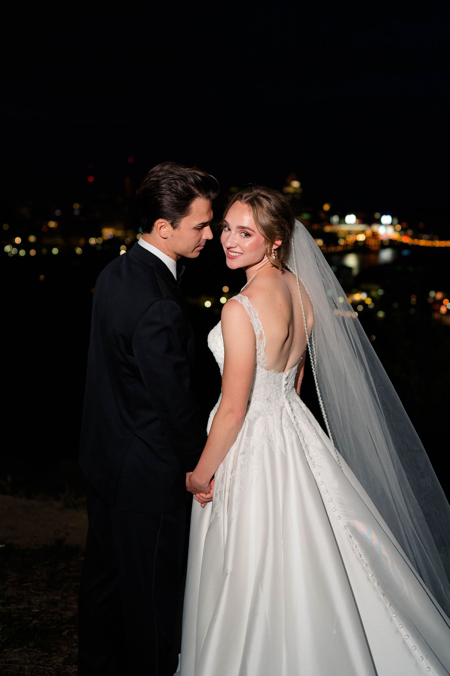 Bride and groom holding hands at night with bokeh city lights in the background during wedding portraits.