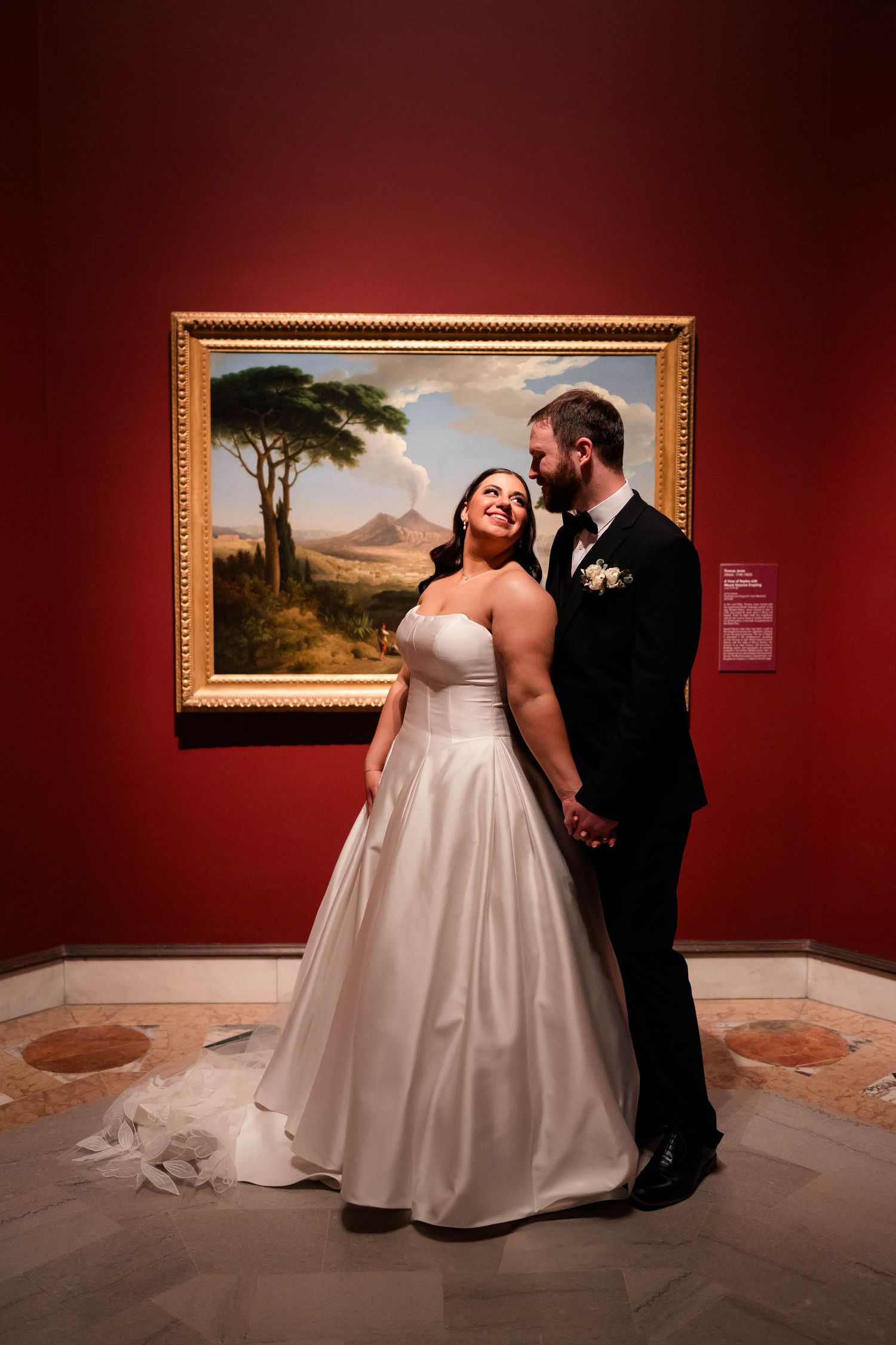 Bride and groom share a romantic moment in front of a gold-framed landscape painting on a red museum wall.