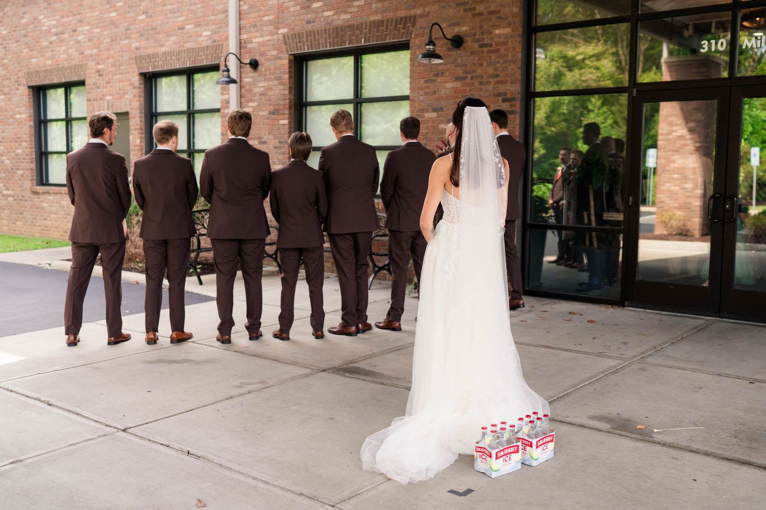 Bride surprising groomsmen in brown suits with beer cases outside brick building.