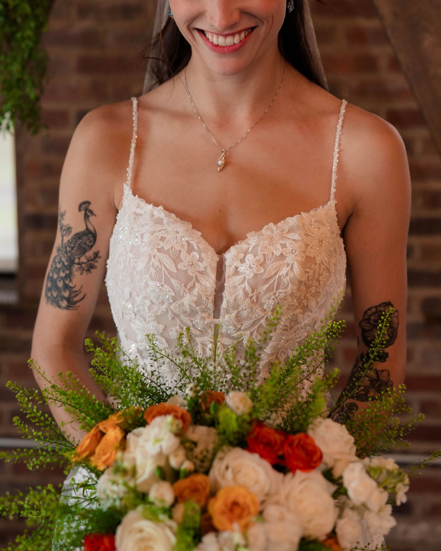 Smiling bride in lace wedding gown holds colorful bouquet of orange and white roses with greenery.
