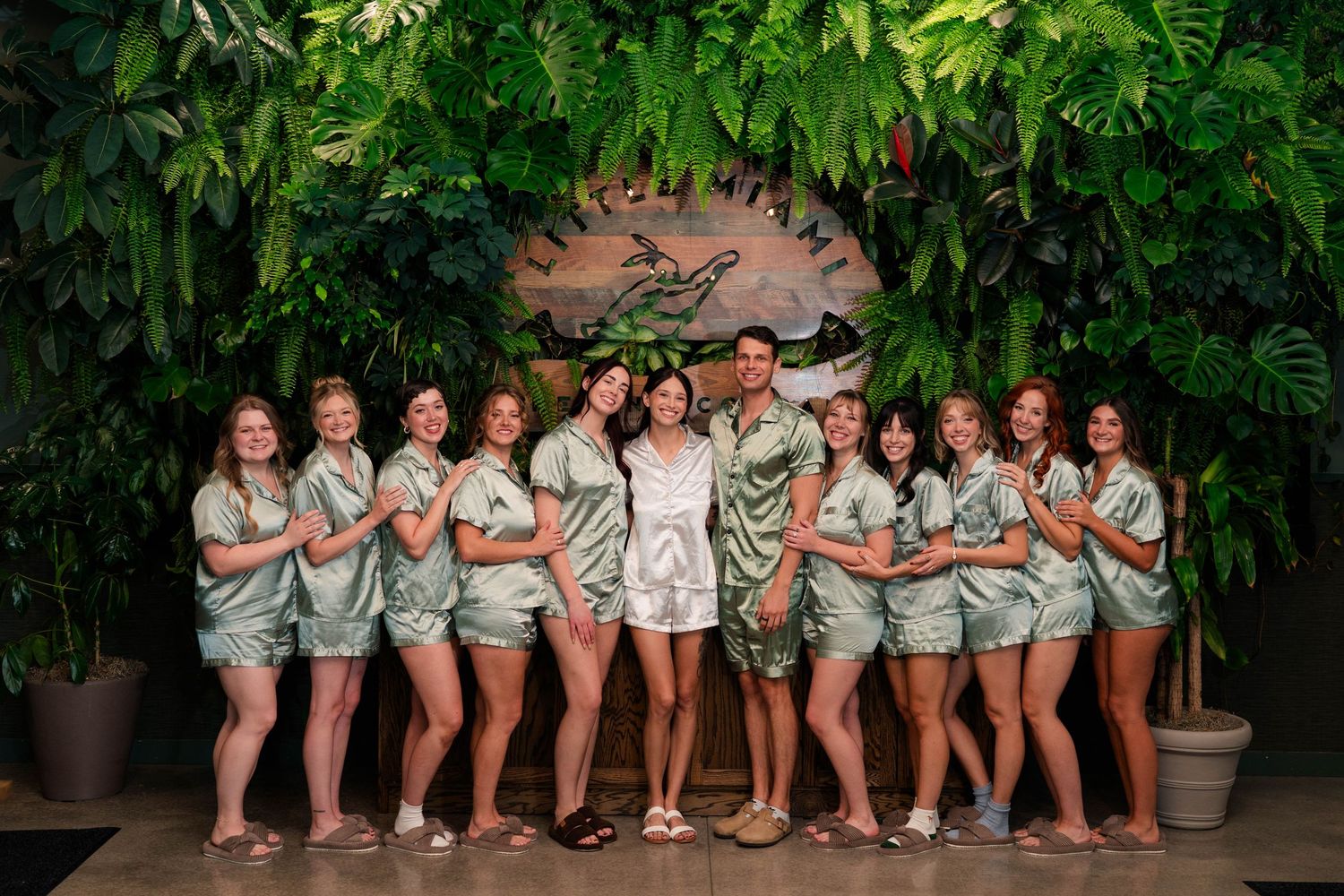 Bride and bridesmaids in matching sage green satin pajamas posing in front of a tropical green wall.