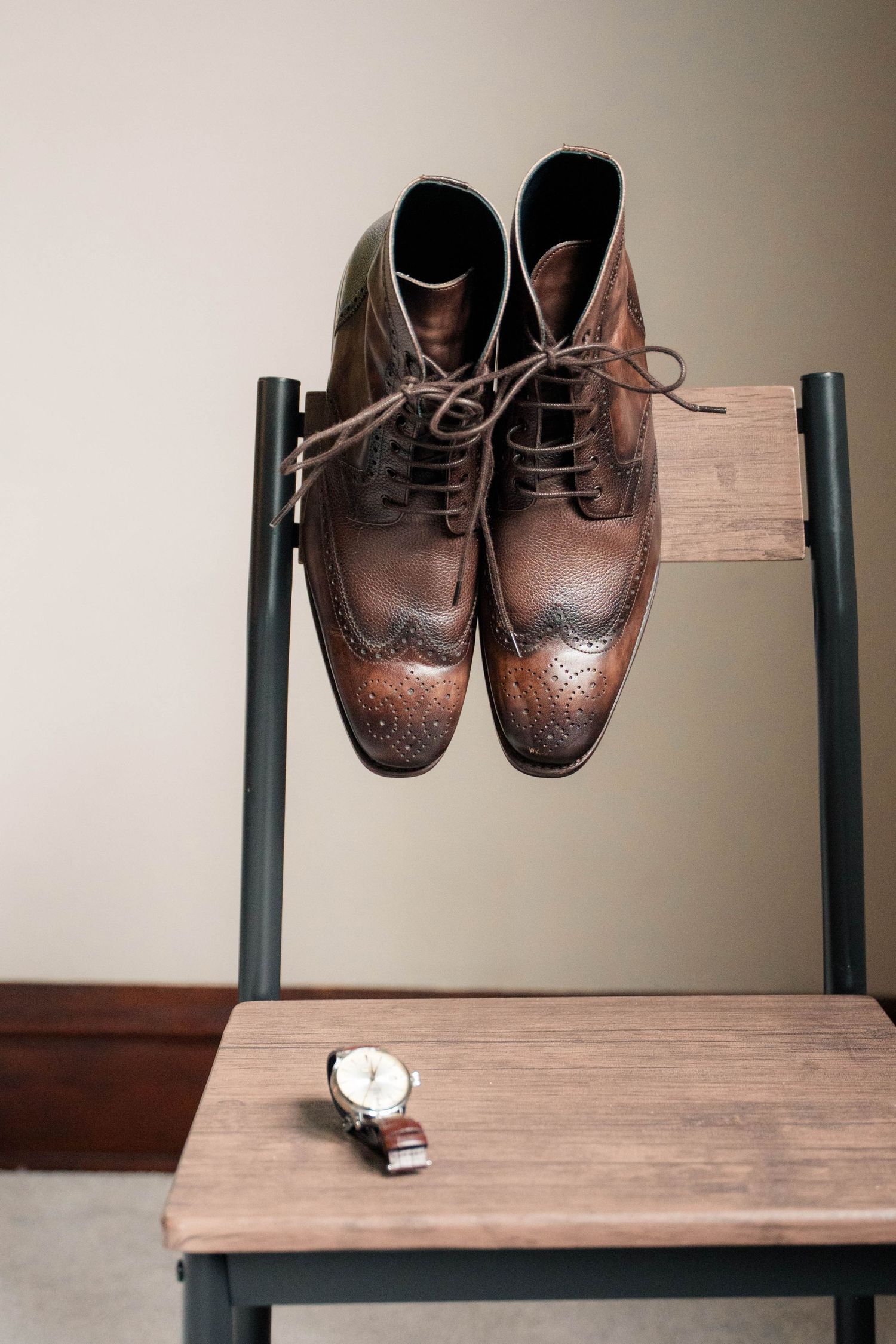 Brown leather brogue boots hanging on a wooden chair back with a wristwatch on the seat below.