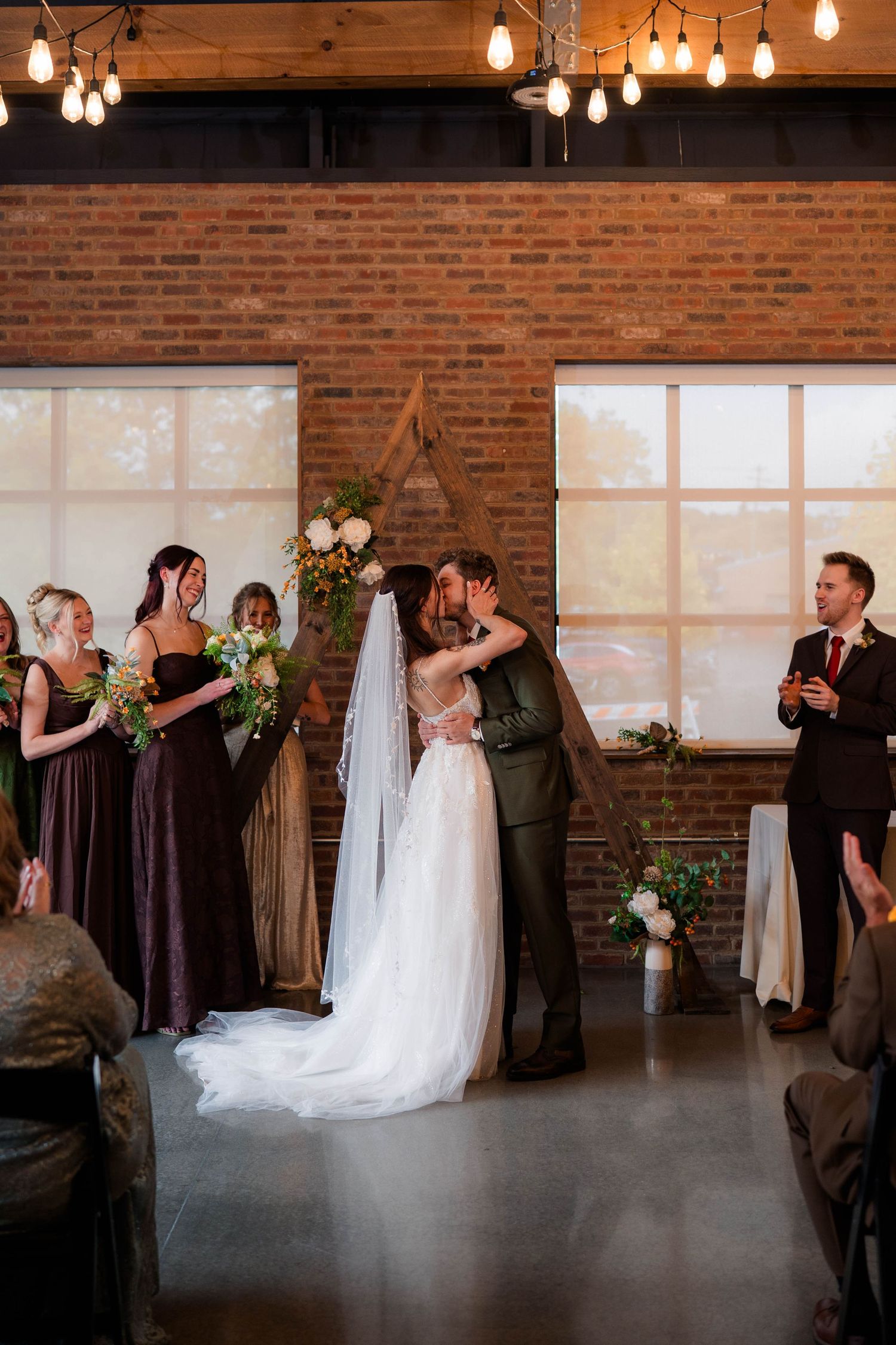 Bride and groom share first kiss at altar in rustic brick venue with Edison bulb lighting.