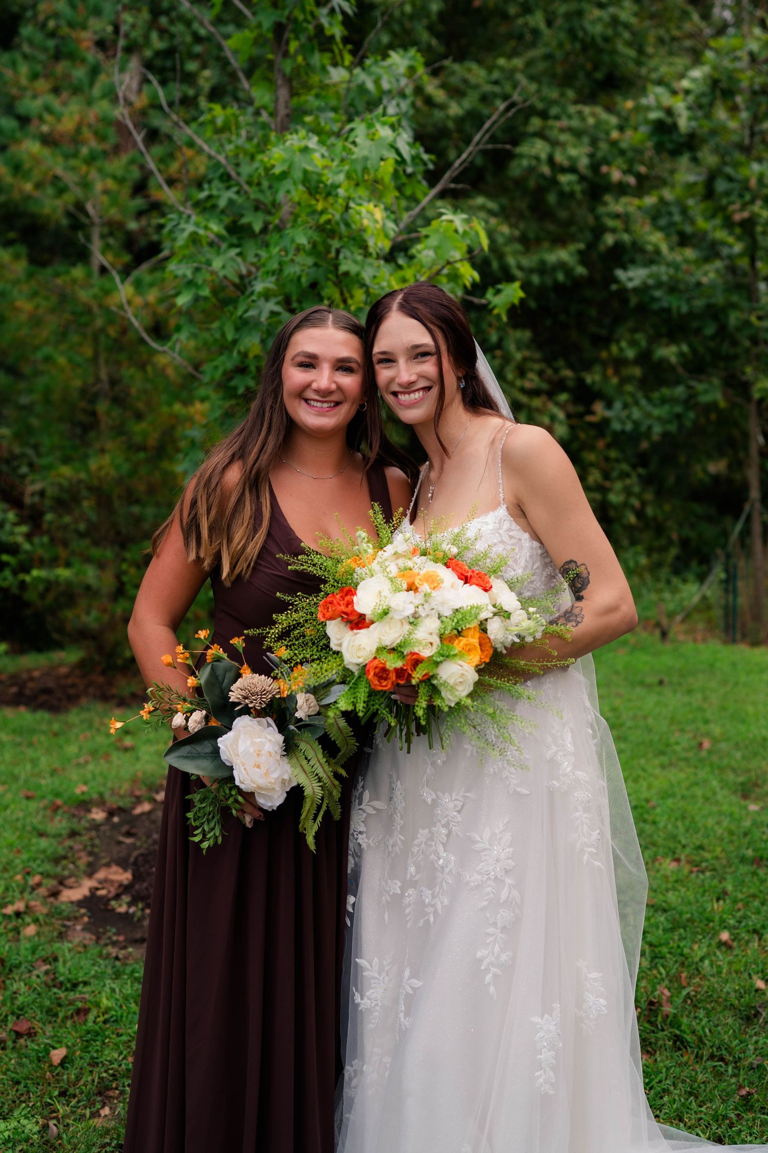 Bride in lace gown and bridesmaid in brown dress holding colorful fall bouquets outdoors.