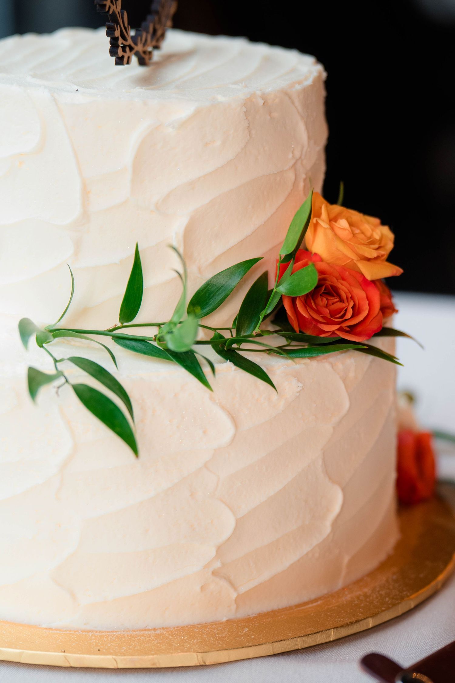 Two-tier white wedding cake with textured frosting, orange roses, and green foliage decoration on a gold base.