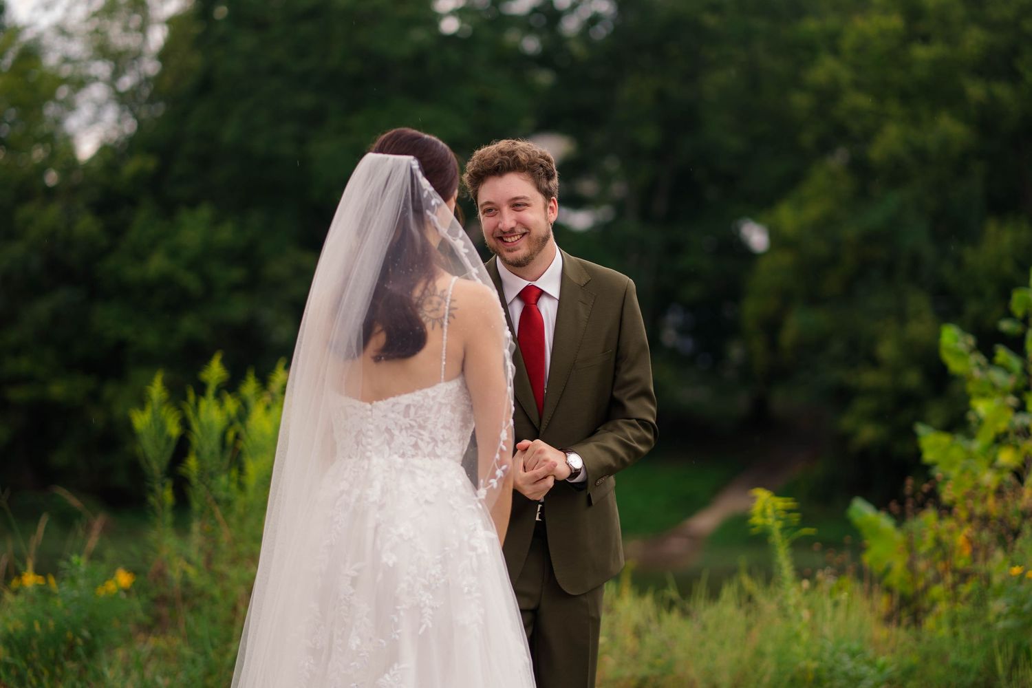 Groom in olive suit smiling at bride during outdoor first look wedding moment.