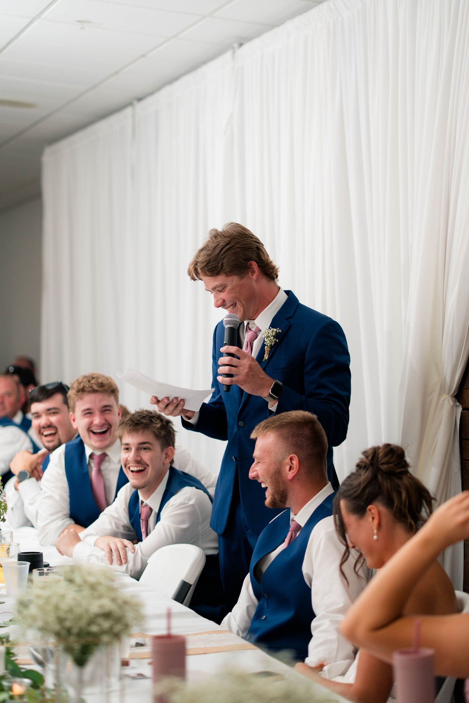 Groomsman in navy suit delivers humorous speech to laughing wedding party at decorated reception dinner table.