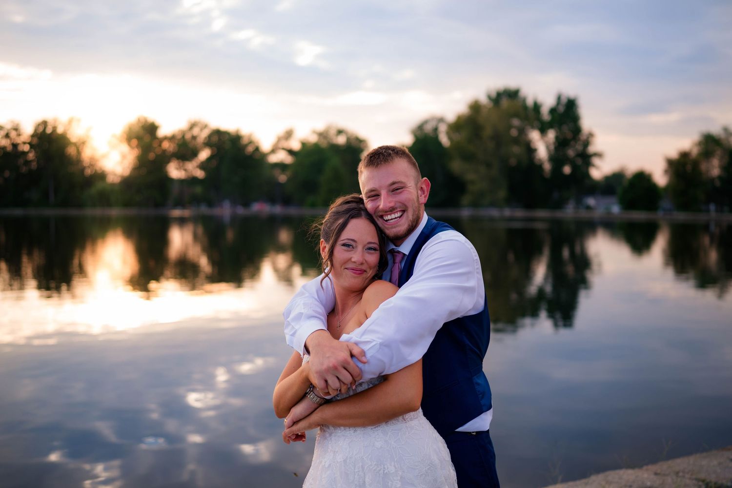Bride and groom embracing by a lake at sunset, smiling joyfully in wedding attire with trees reflected in water.