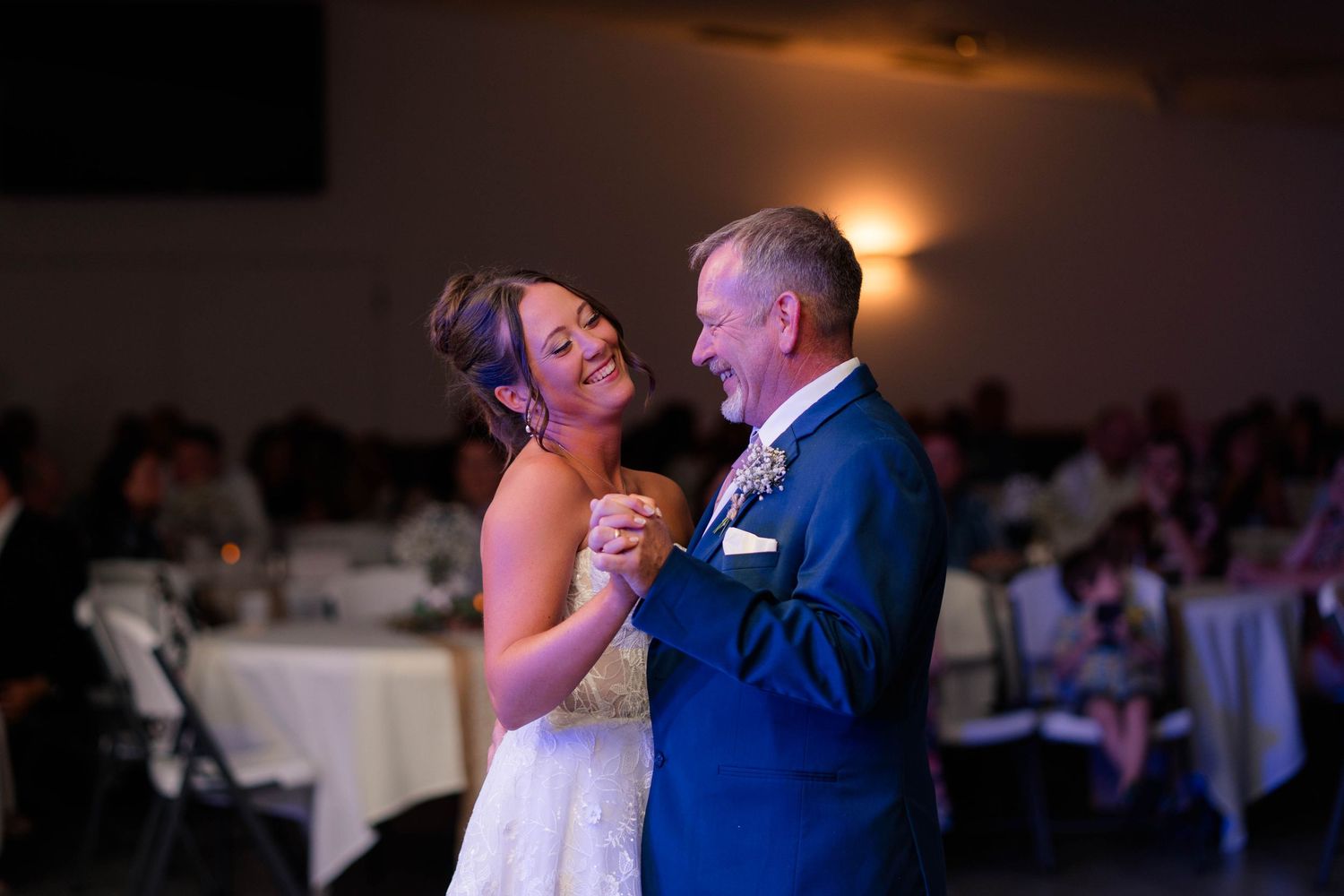 Bride shares a joyful father-daughter dance at elegant wedding reception in blue-lit venue.
