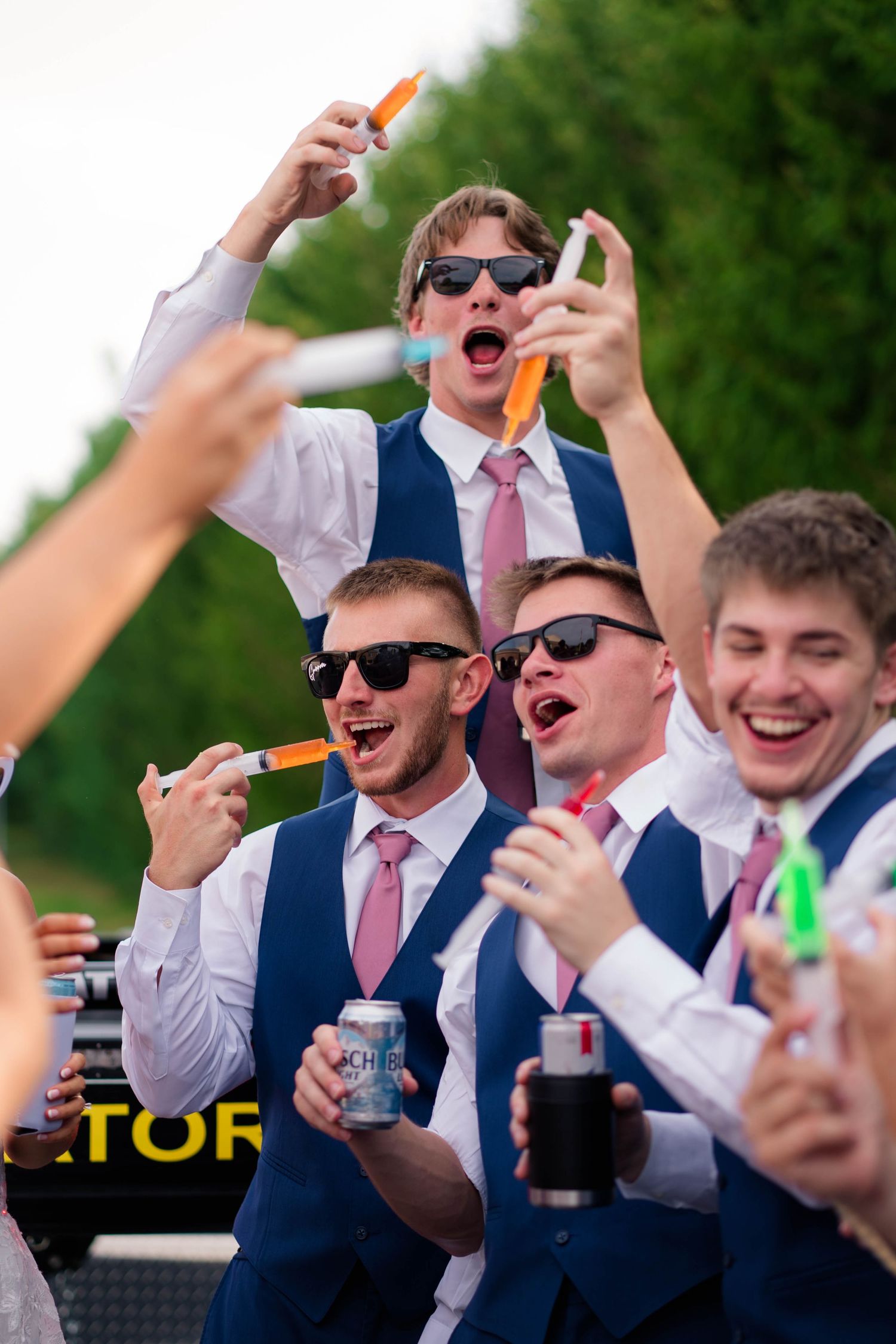 Groomsmen in navy suits and pink ties celebrate with drinks and syringes outdoors.