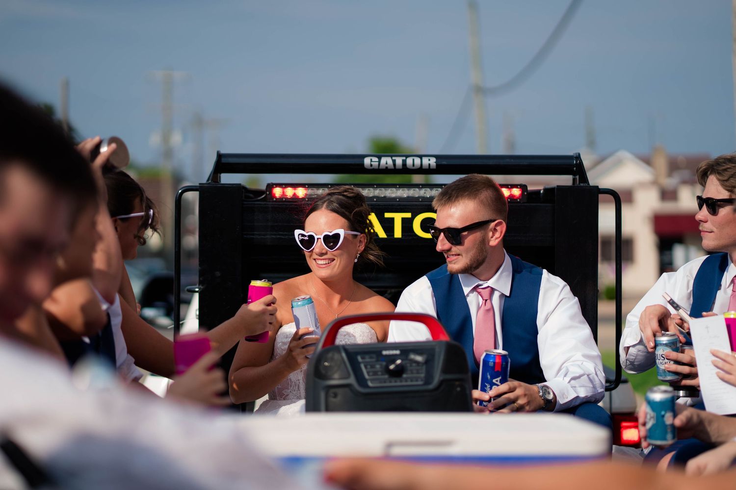 Bride in white dress and groom in navy suit riding in a Gator vehicle at wedding reception.