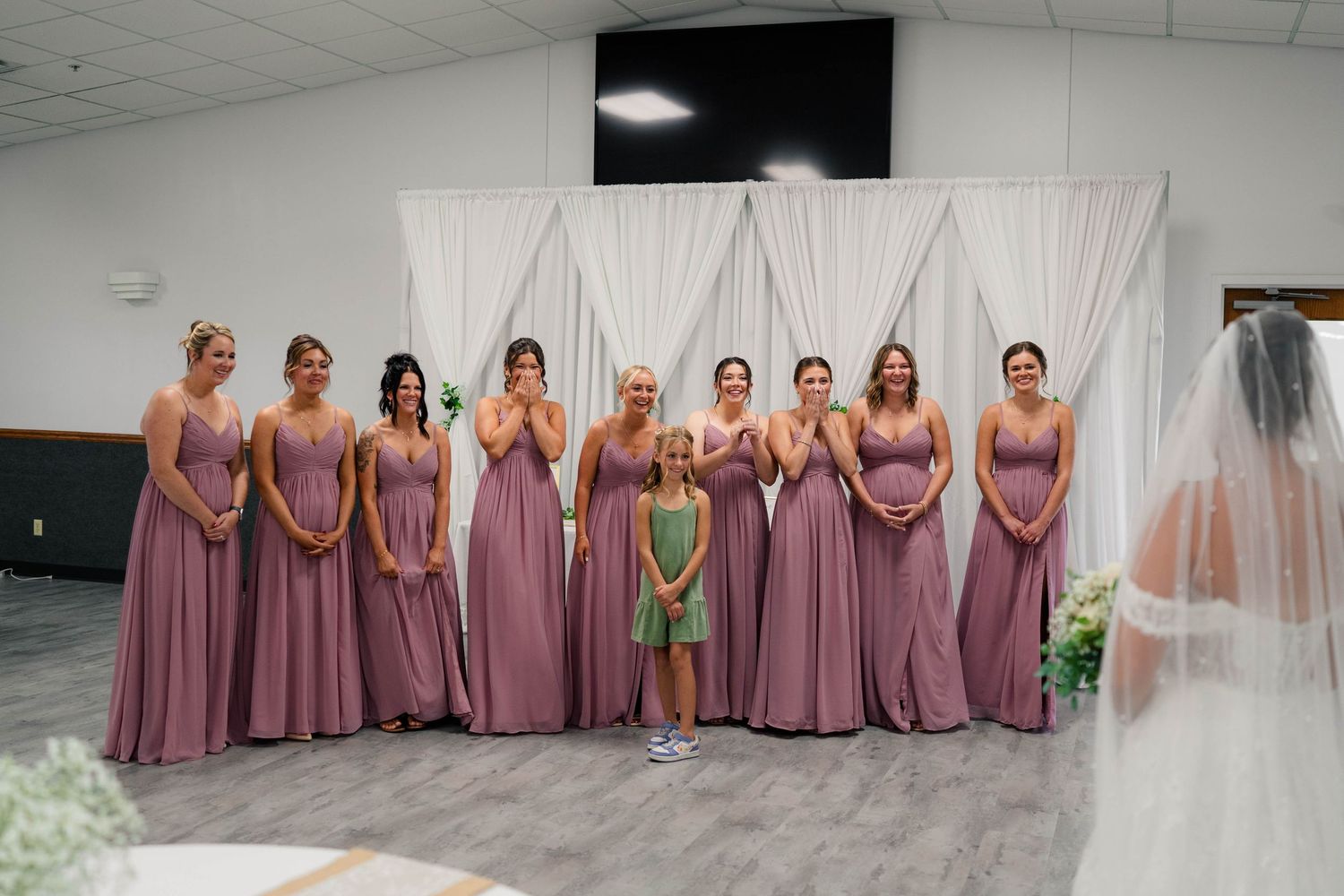 Bridesmaids in mauve dresses stand smiling beside bride in white gown and veil at wedding ceremony.