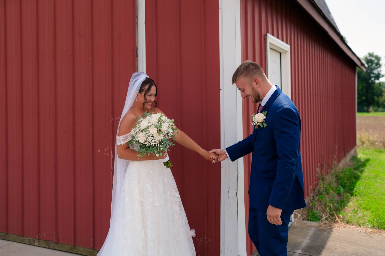 Groom reaching for brides hand as she holds bouquet during first look at red barn wedding venue.