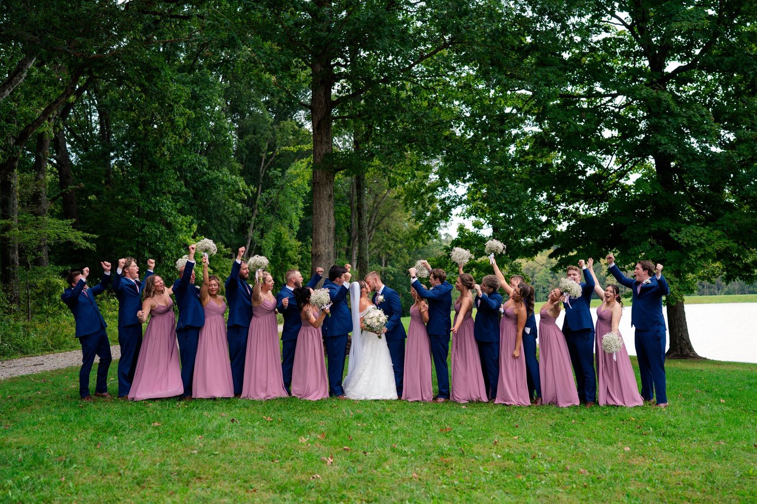 Large wedding party in navy suits and dusty rose gowns celebrate on a lush green lawn.