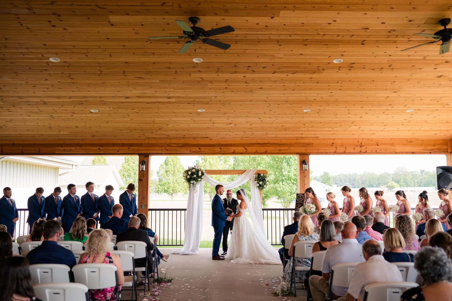 Outdoor wedding ceremony under wooden pavilion with guests seated and couple at floral arch altar.