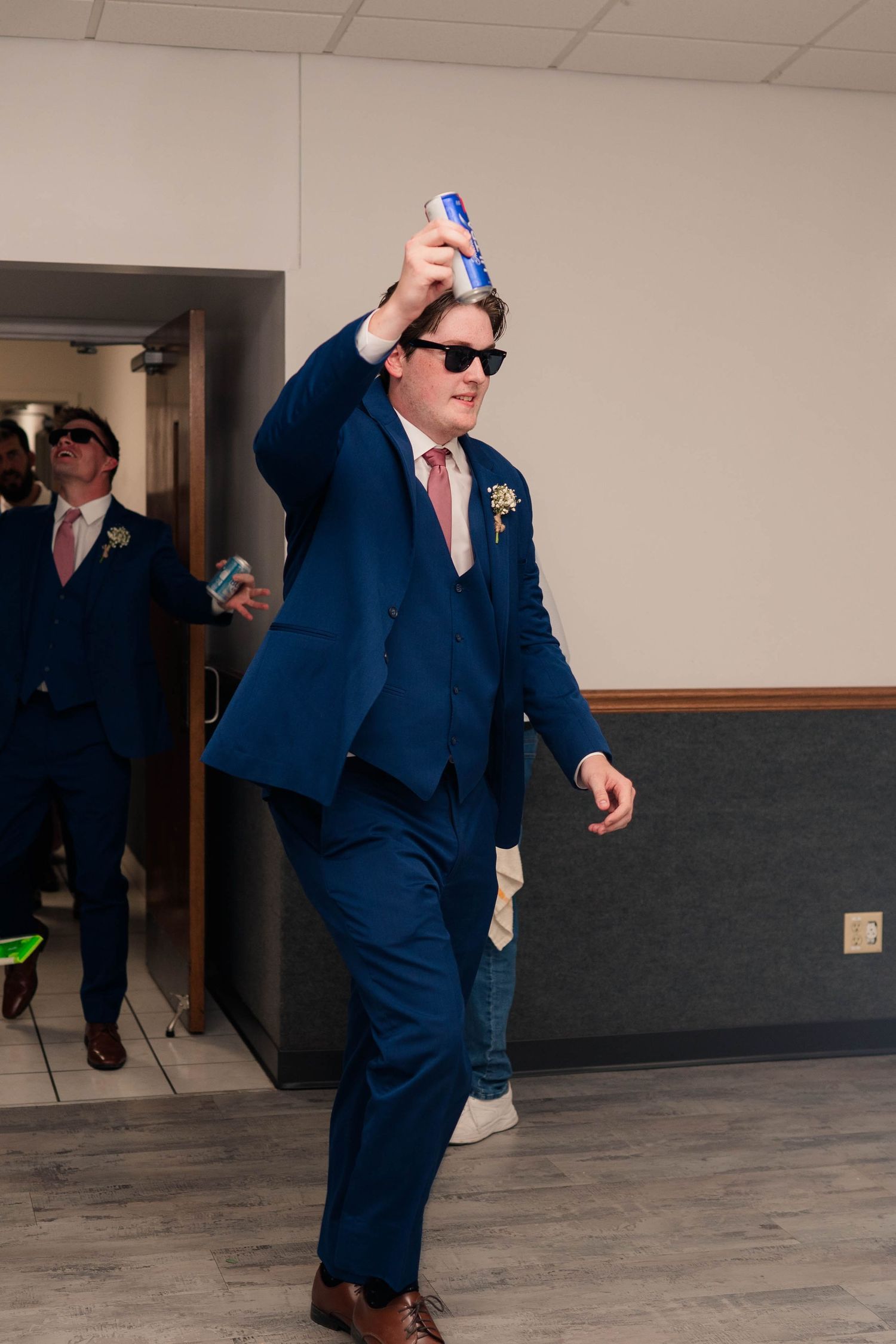 Groomsman in navy suit and sunglasses raises beer can while making grand entrance at wedding reception.