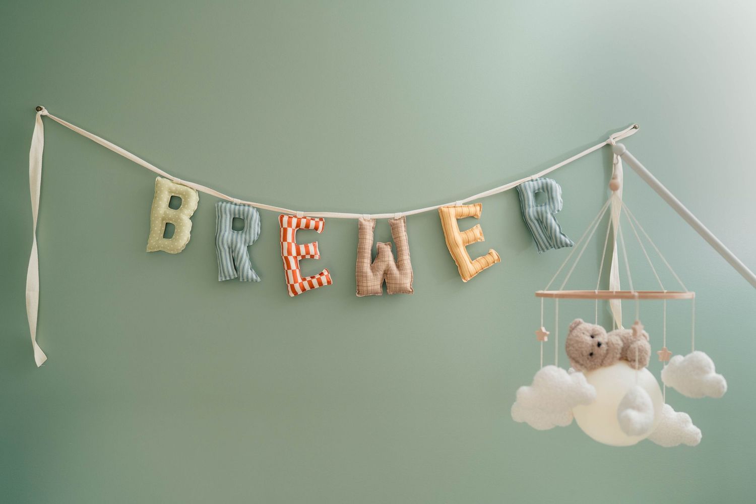 Colorful fabric letter banner spelling BREWER with a teddy bear cloud mobile on a sage green wall.