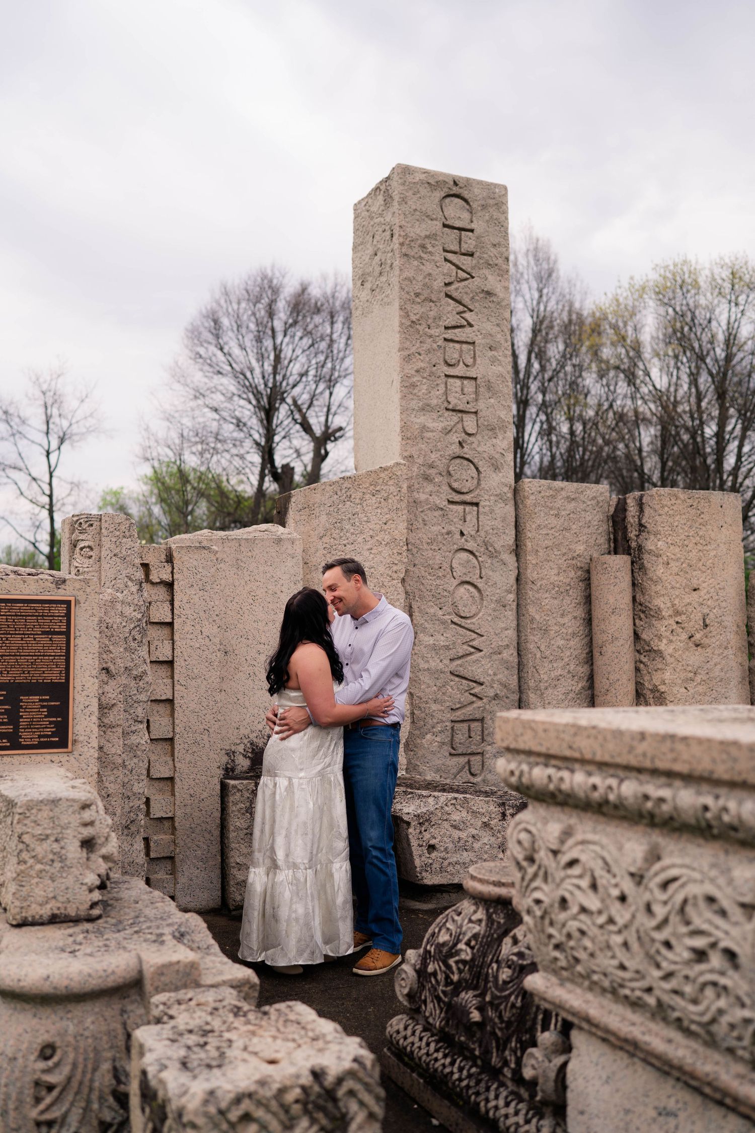 Couple embracing romantically among stone ruins with Chamber of Commerce pillar in background.