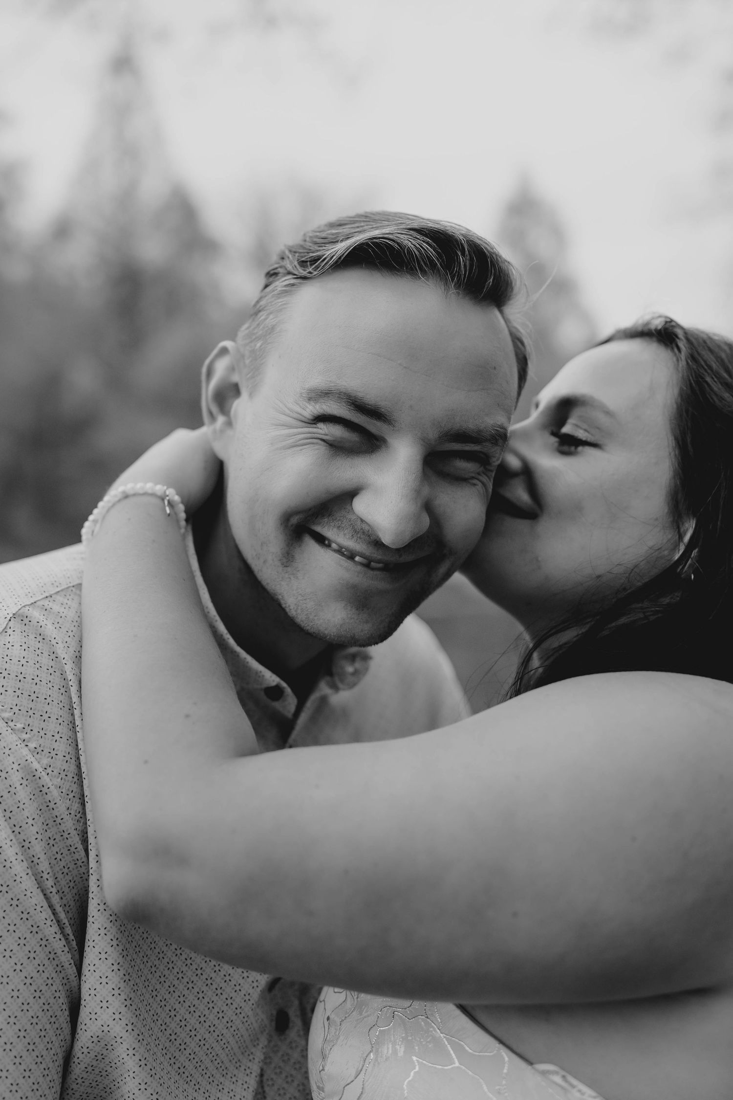 Black and white photo of a smiling man being kissed on the cheek by his partner outdoors.