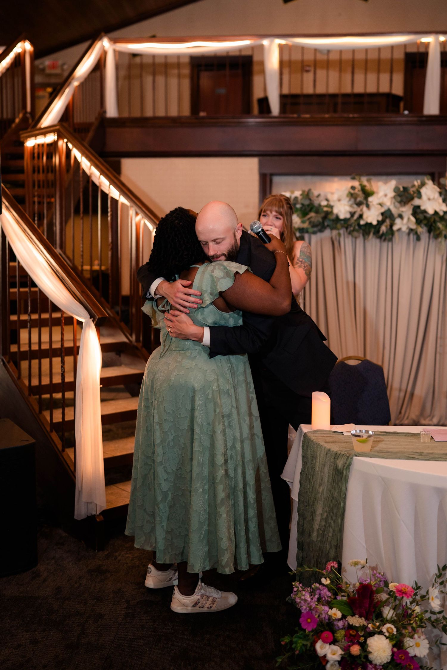 Emotional wedding reception hug between guests near staircase with floral decorations and warm lighting.