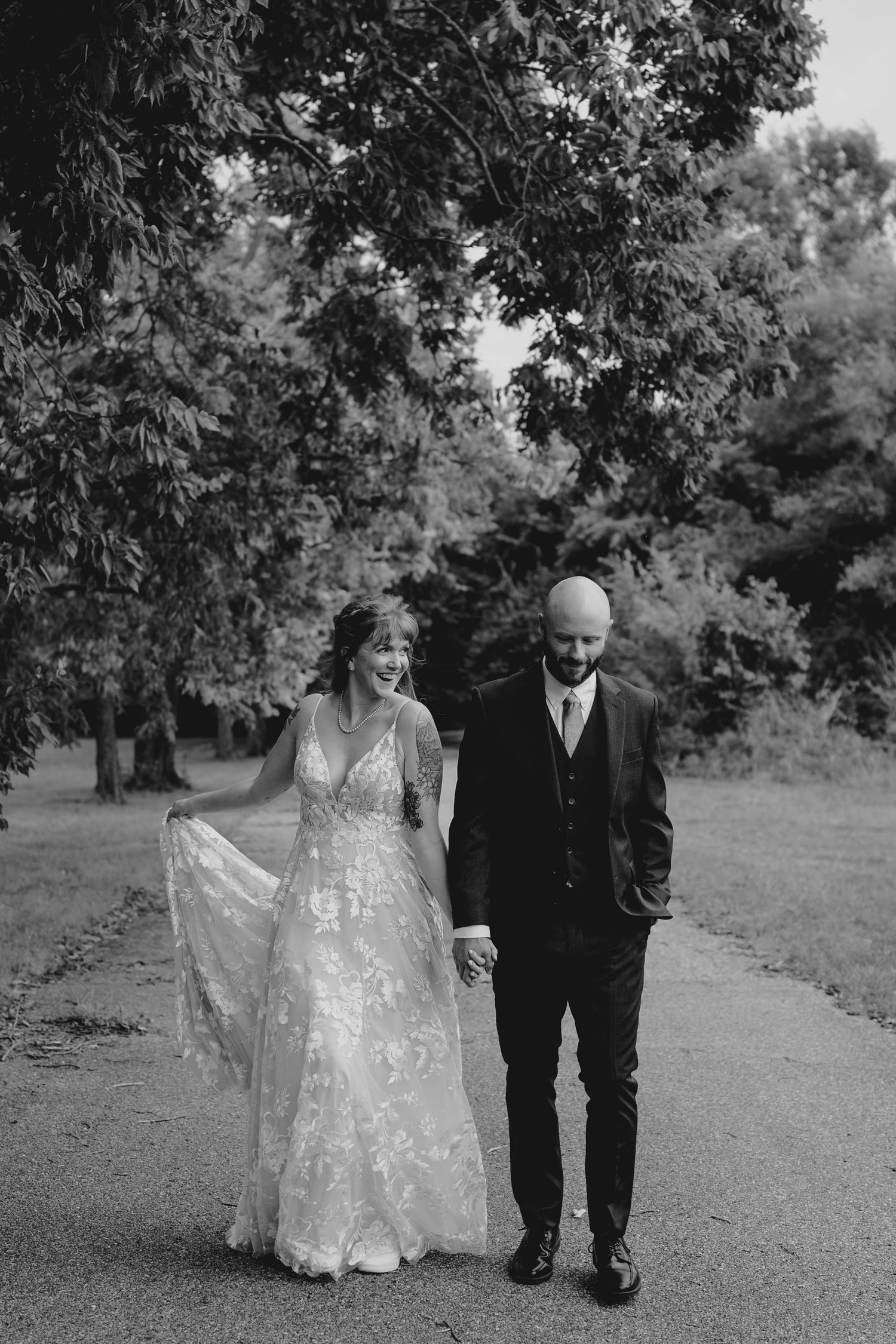 A black and white photo of a laughing bride and groom walking hand in hand down a tree-lined path.
